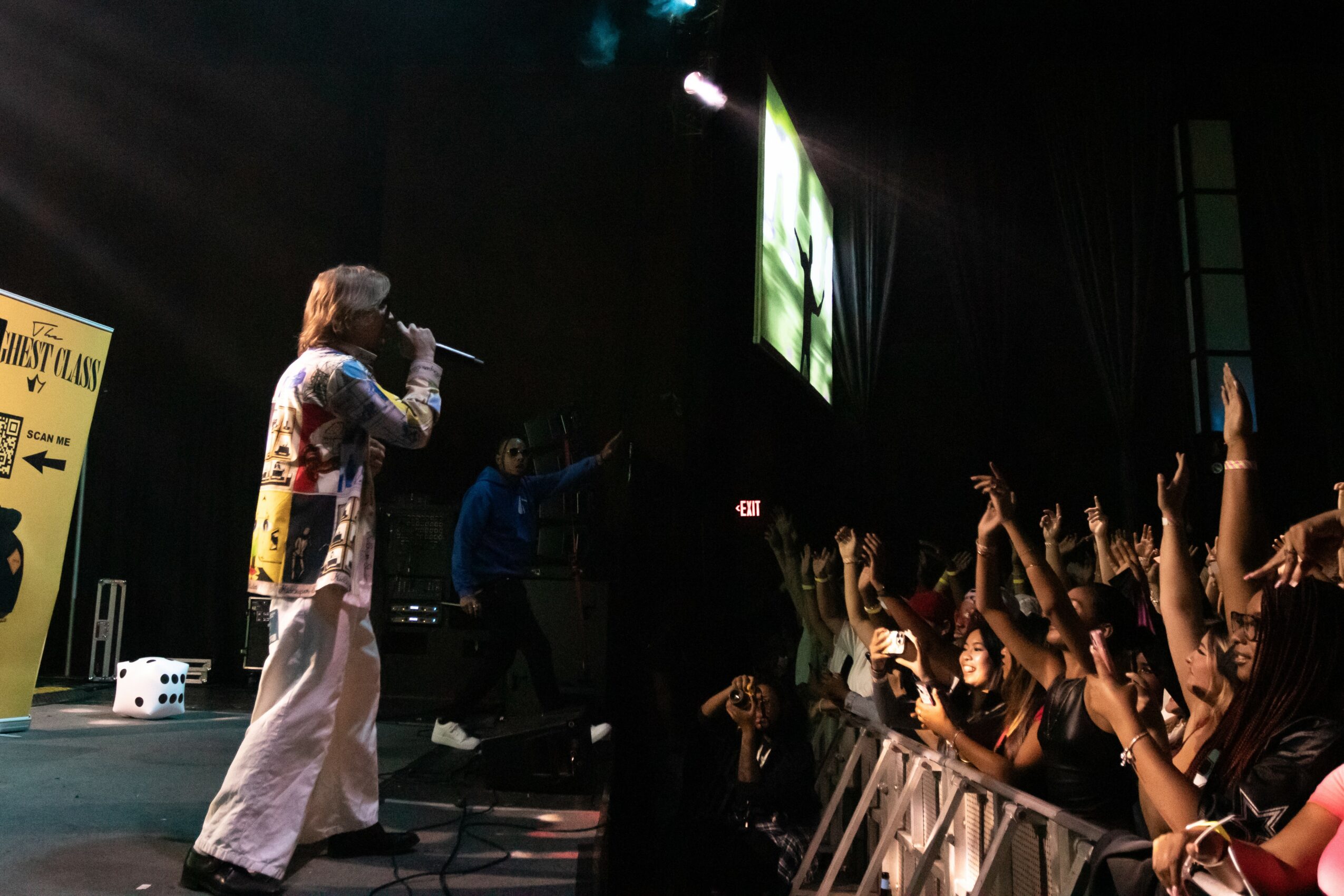 Performer on stage singing to an audience with hands raised, stage lights, and a large screen behind.
