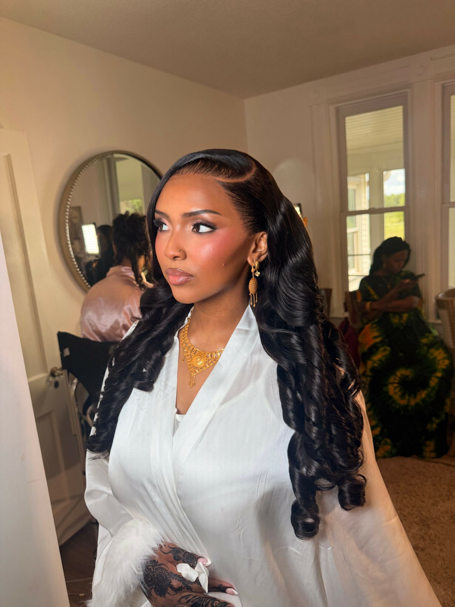 Woman with long, curled hair wearing a white top and gold jewelry, standing indoors near a mirror and window.