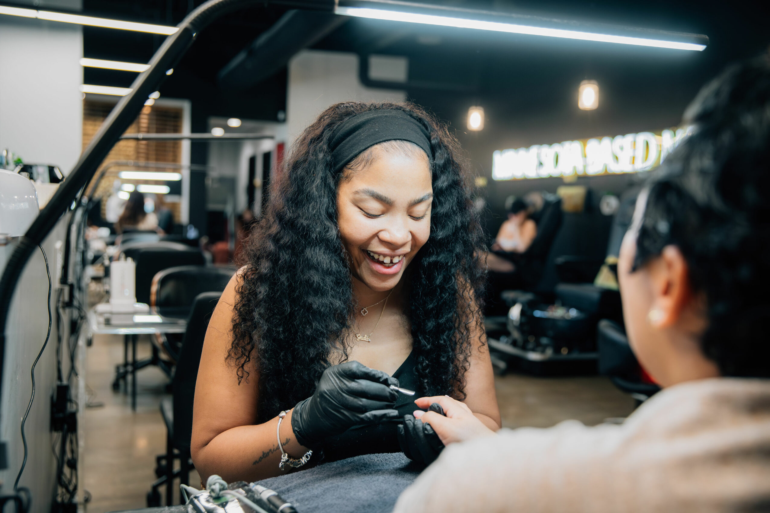 Smiling woman with curly hair and black headband, wearing black gloves, in a modern indoor setting with people and equipment.