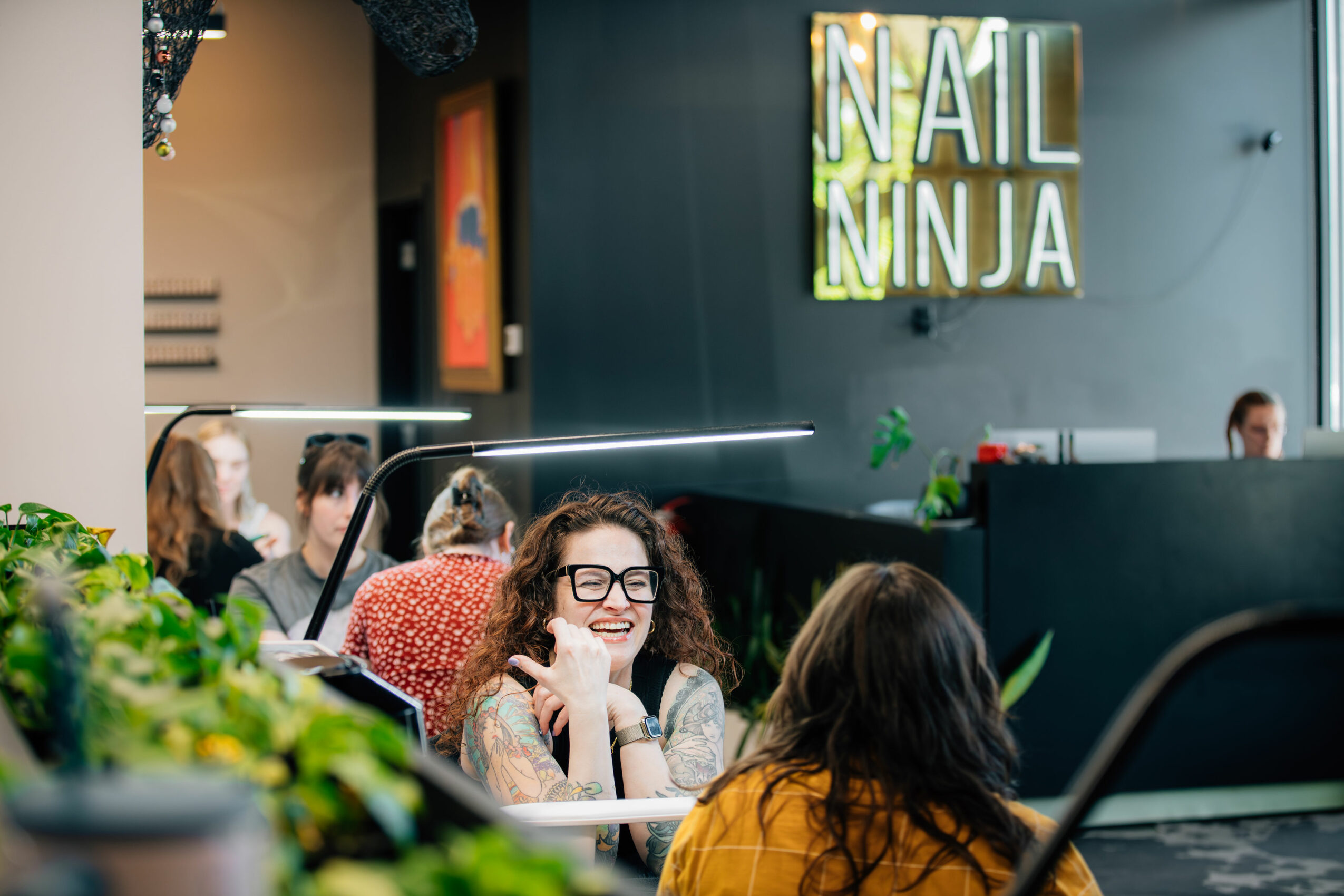 People sitting at a nail salon, with a sign reading 'NAIL NINJA' on the wall, woman smiling, others in background.