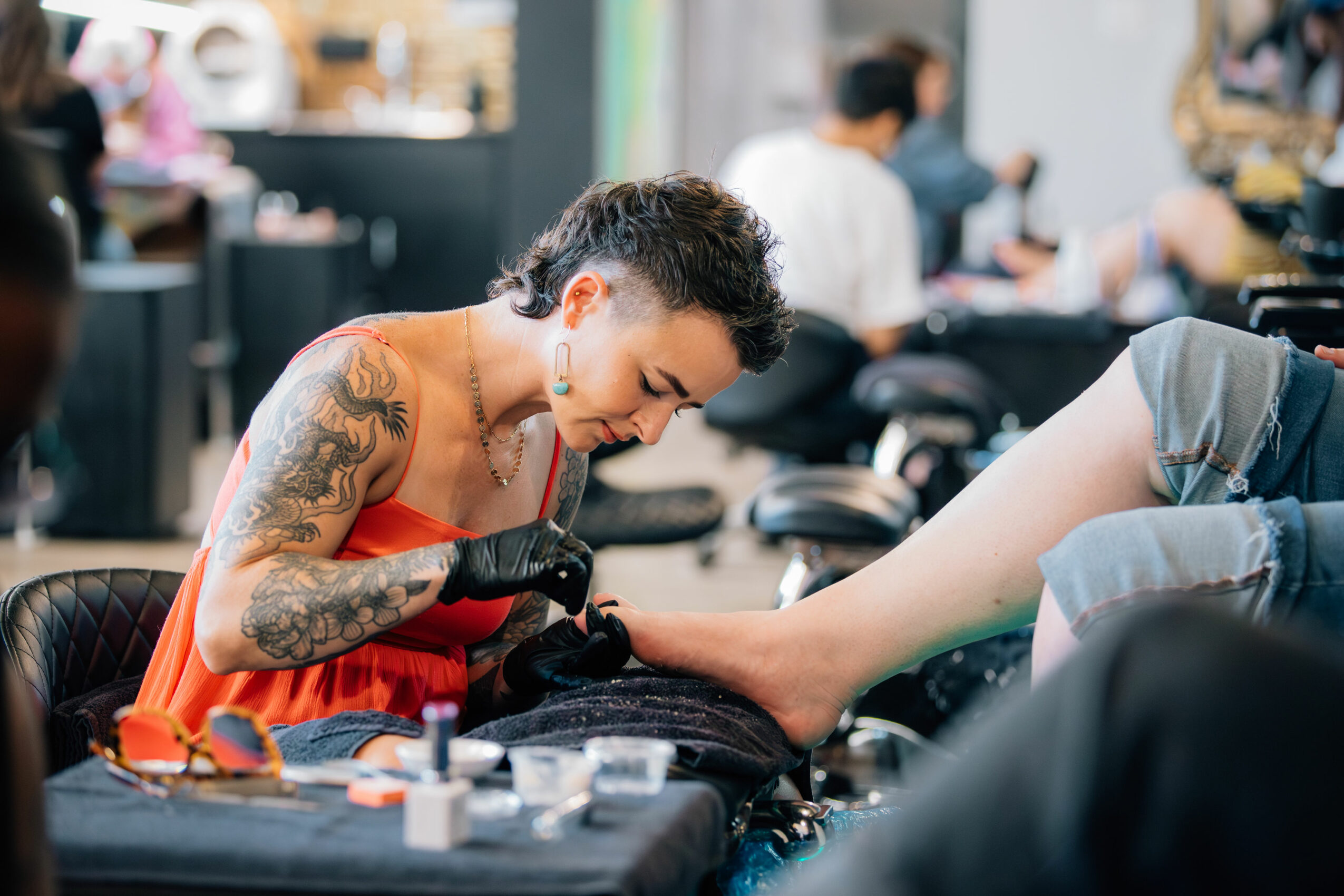 Tattooed woman receiving a pedicure in a salon, with other clients and staff in the background.
