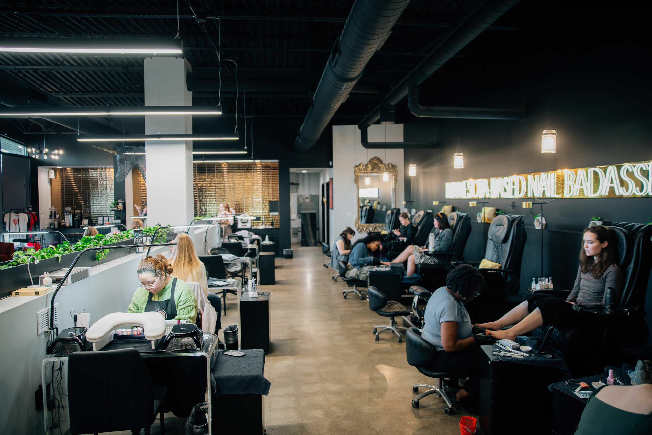 People sitting in chairs at nail salon stations, receiving manicure treatments, with a modern interior and neon sign in background.