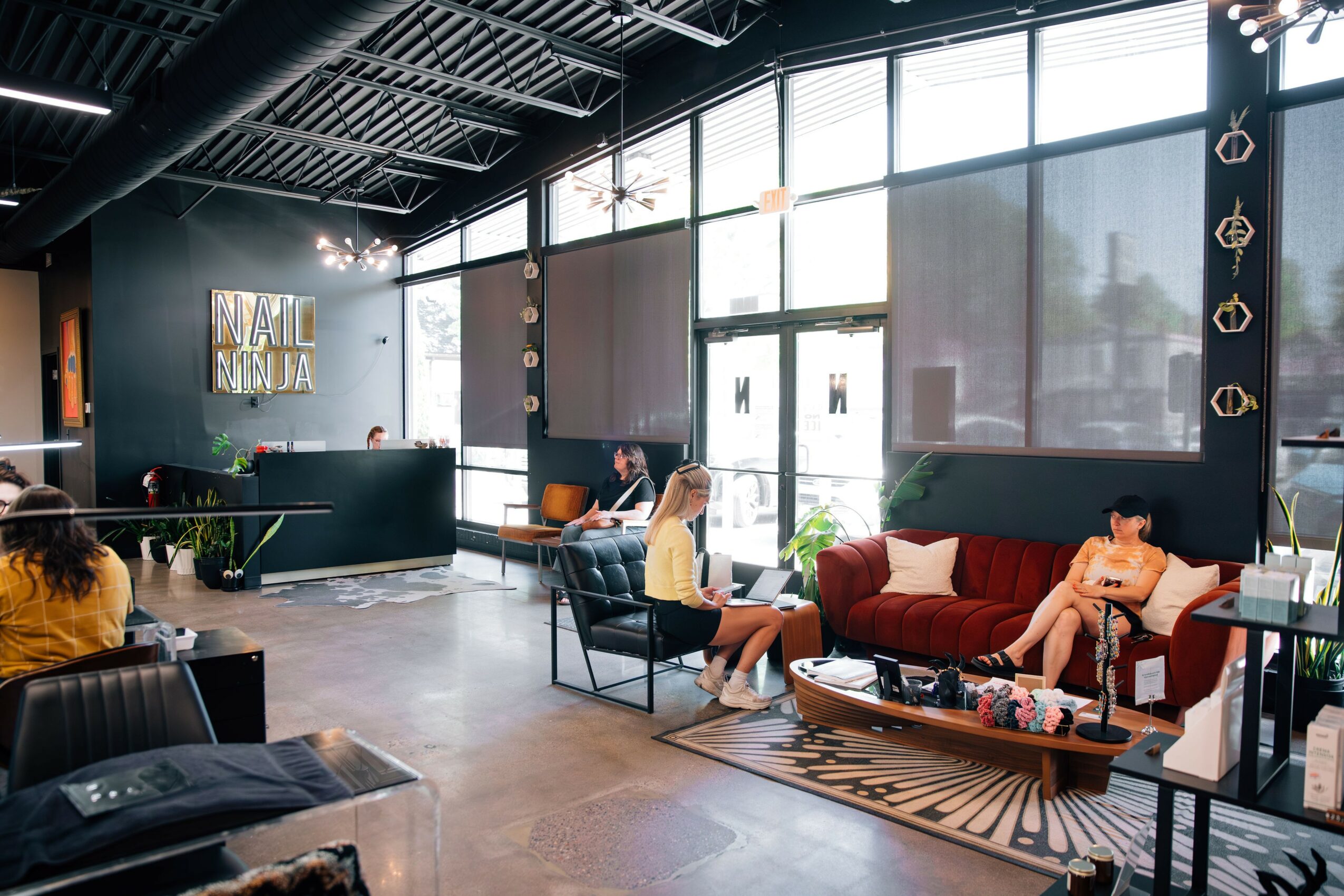 Modern lobby with large windows, seating area with red sofa, people sitting and talking, decorative wall elements, and a reception desk.