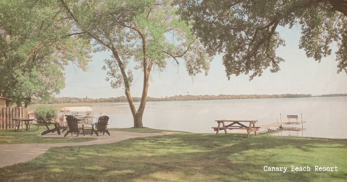Scenic lakeside view with trees, benches, and a picnic table, under a partly cloudy sky.