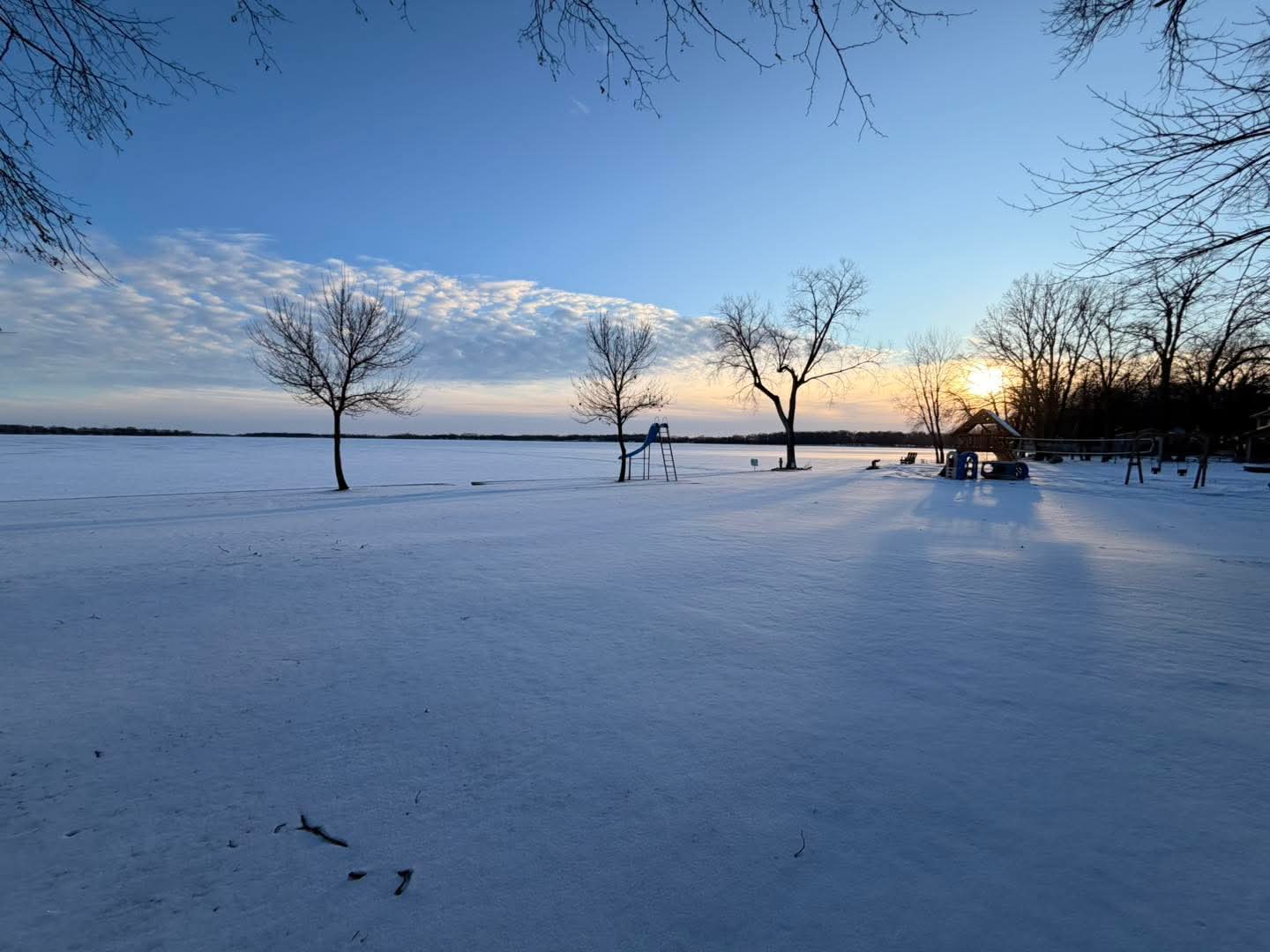 Snow-covered landscape with leafless trees, a fence, and a setting sun on the horizon, under a partly cloudy sky.