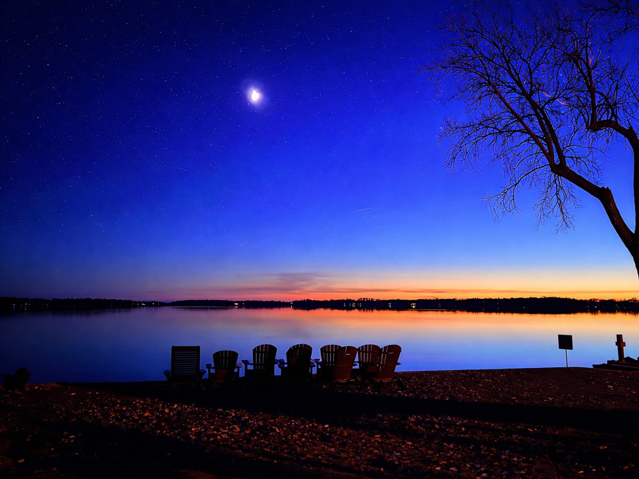 Night scene with a bright moon, a tree on the right, and a calm lake reflecting the sky, with chairs on the shore.