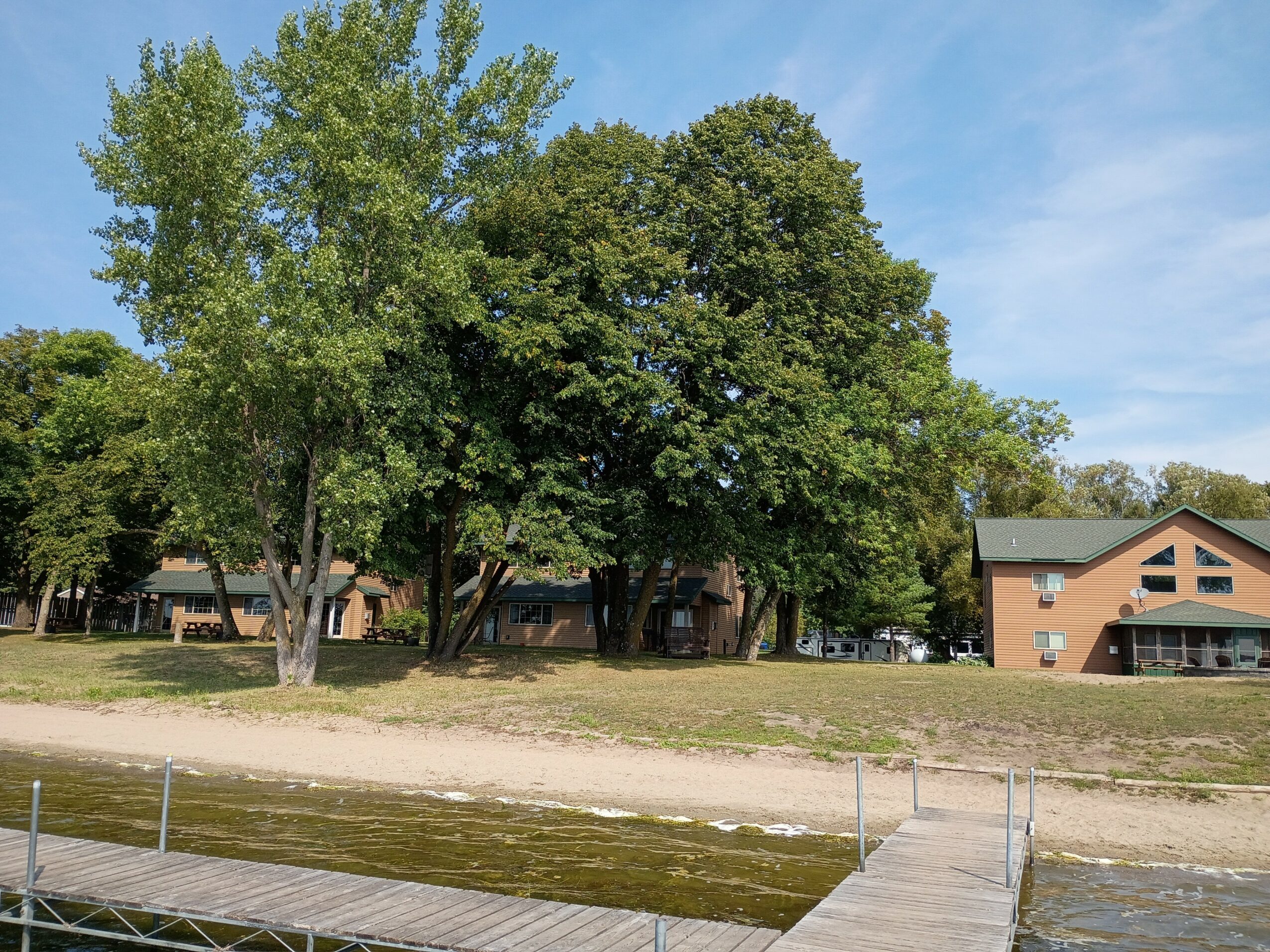 Trees and buildings near a body of water with a dock, under a blue sky with some clouds.