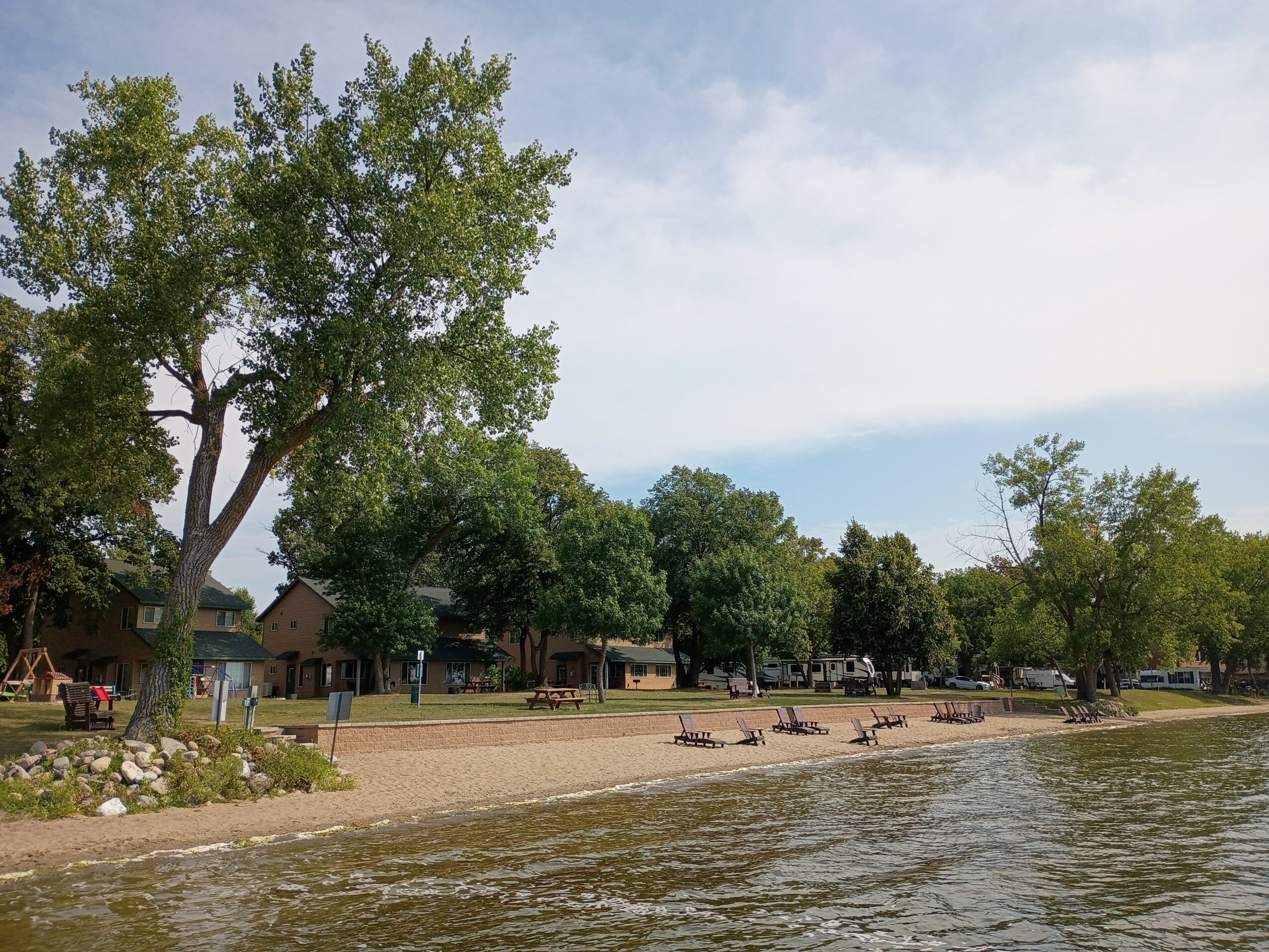 Beach with trees, benches, and water, under a partly cloudy sky.