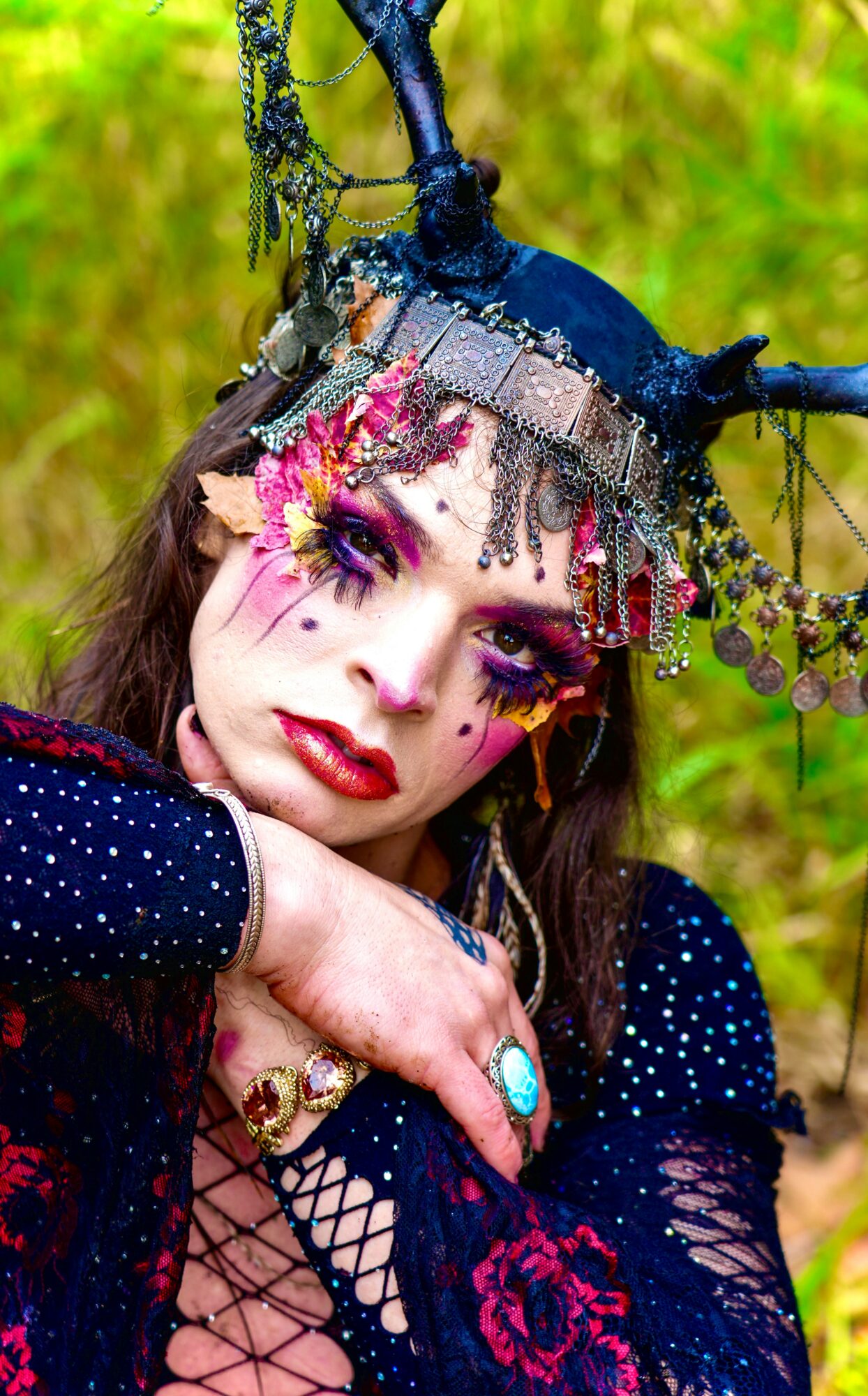 Woman with elaborate headpiece and makeup, wearing dark clothing with lace, outdoors with green background.