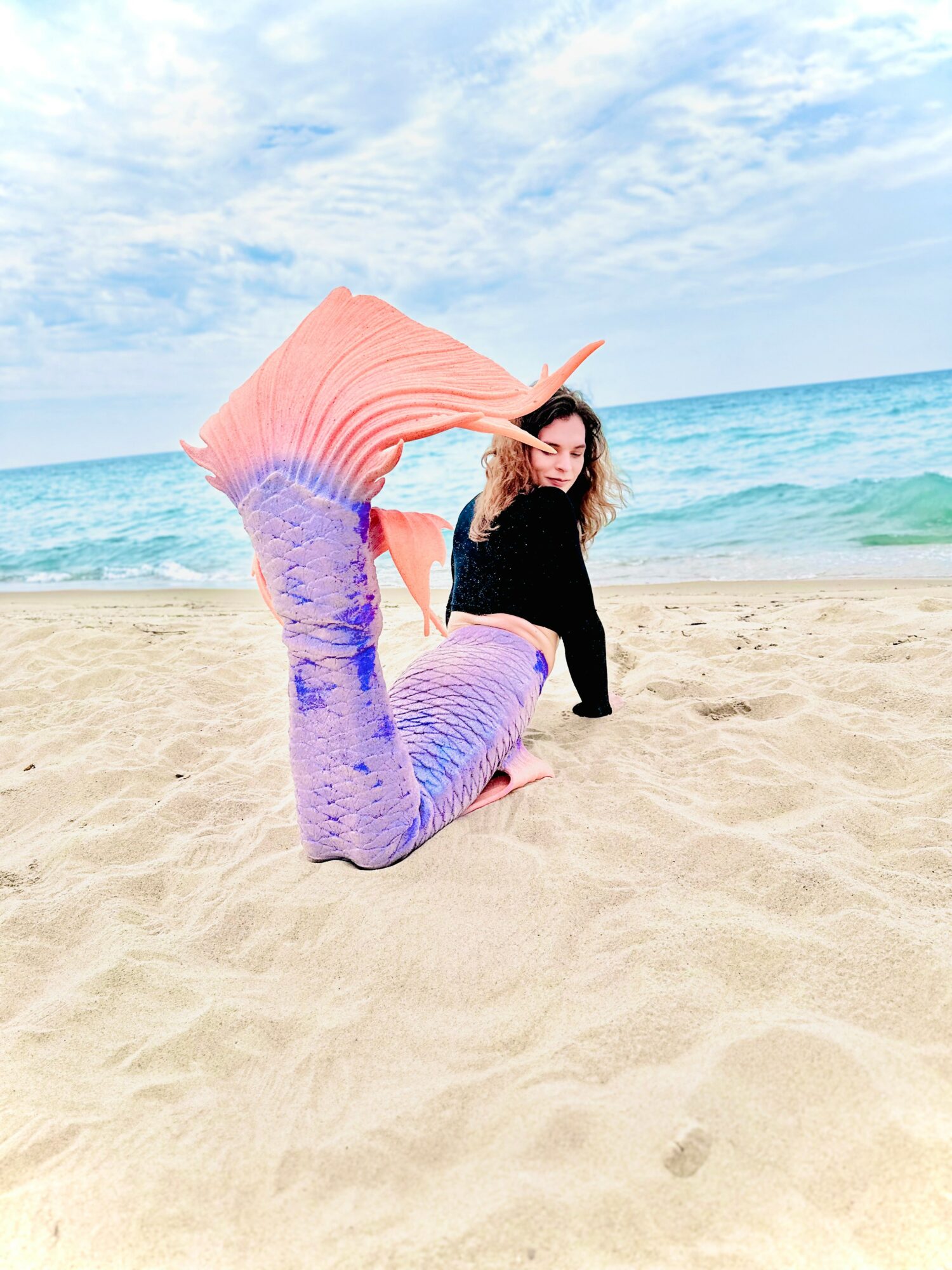 Person with a mermaid tail on a sandy beach, ocean and sky in background, waving hair, smiling, wearing black top.