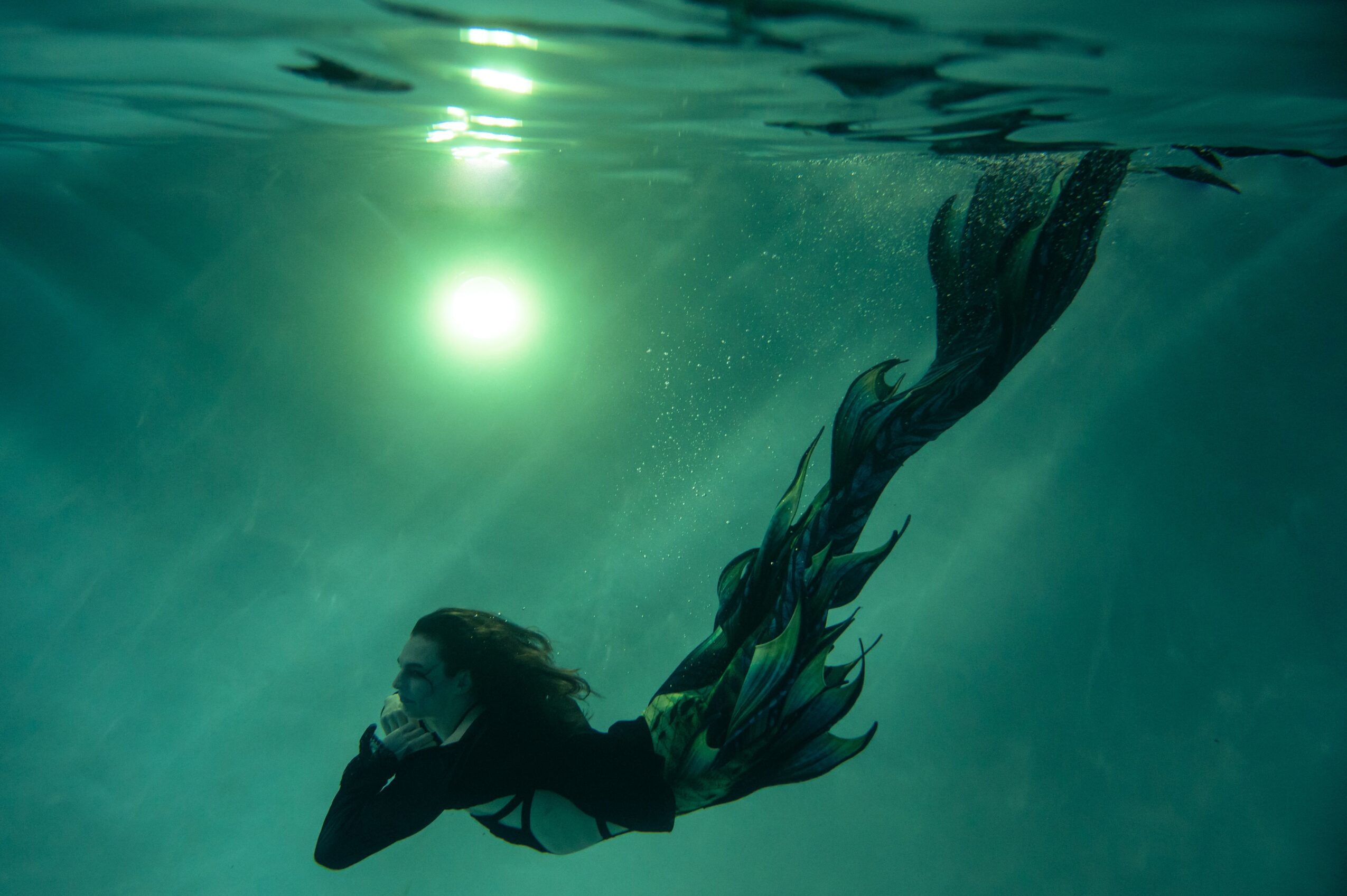 Person swimming underwater with sunlight above, long hair flowing, wearing a wetsuit, in clear water