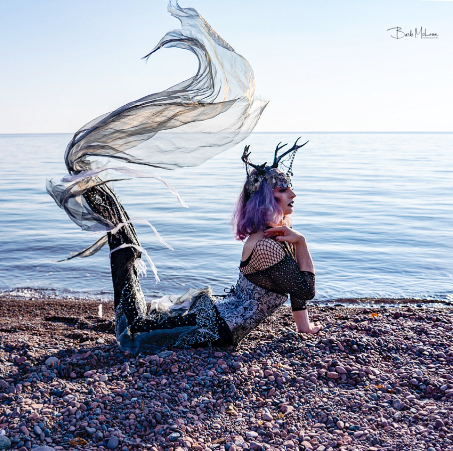 Person with purple hair and antler headband lying on rocky beach near water, with a flowing fabric or sculpture behind them.