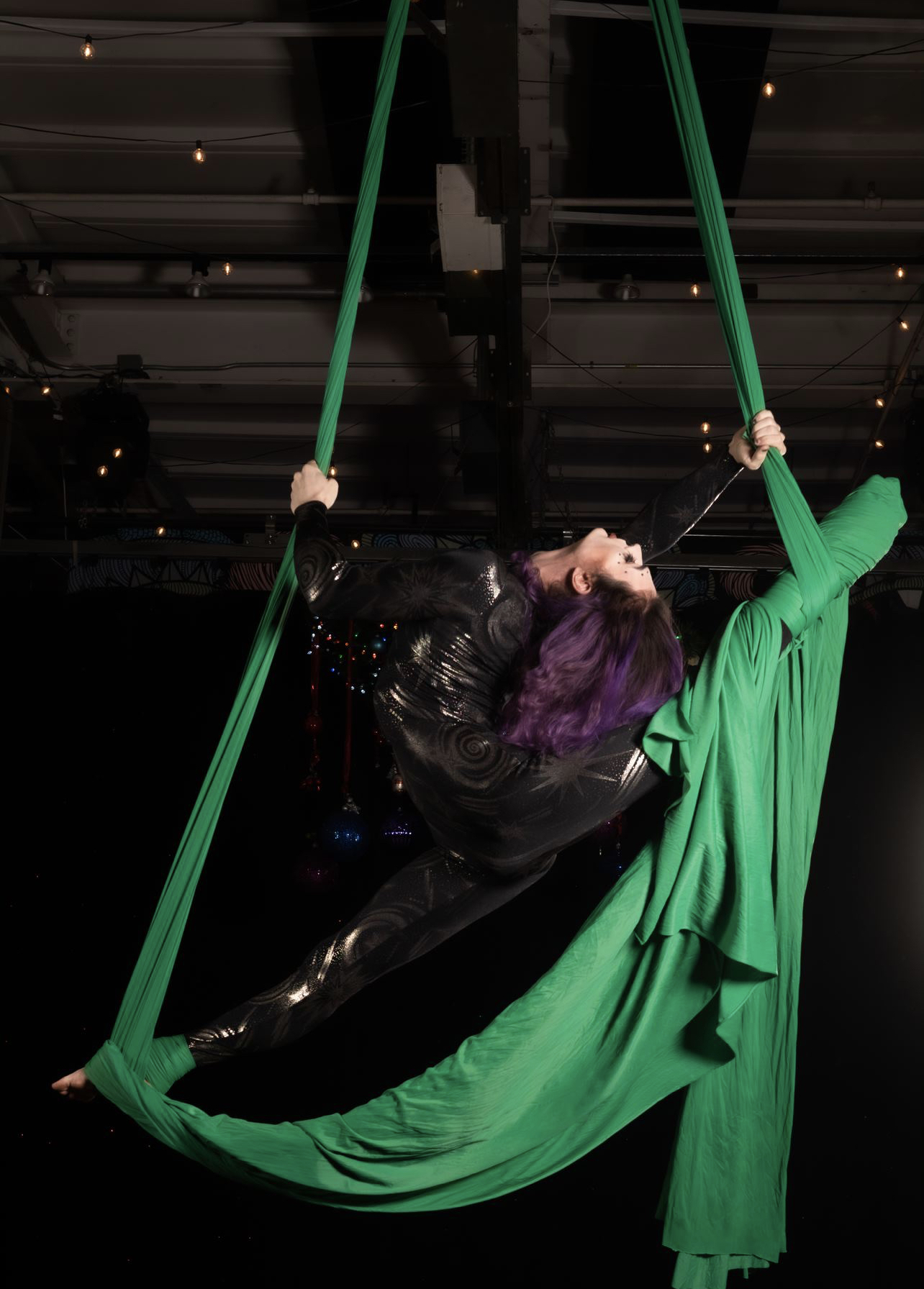 Person with purple hair performing aerial silk act on green fabric in a dark indoor setting.