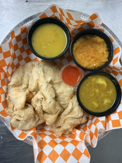 Fried fish with three small bowls of sauces and a cup of yellow liquid on orange checkered paper.