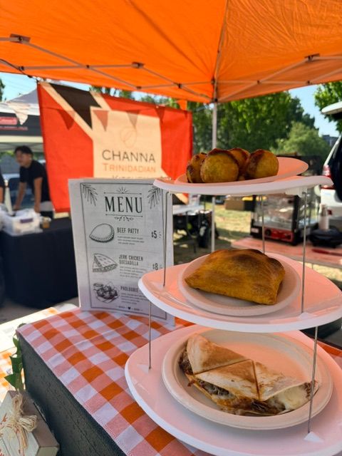 Outdoor food stand with plates of cake and a menu sign, orange umbrella in background, checkered tablecloth, people in distance.