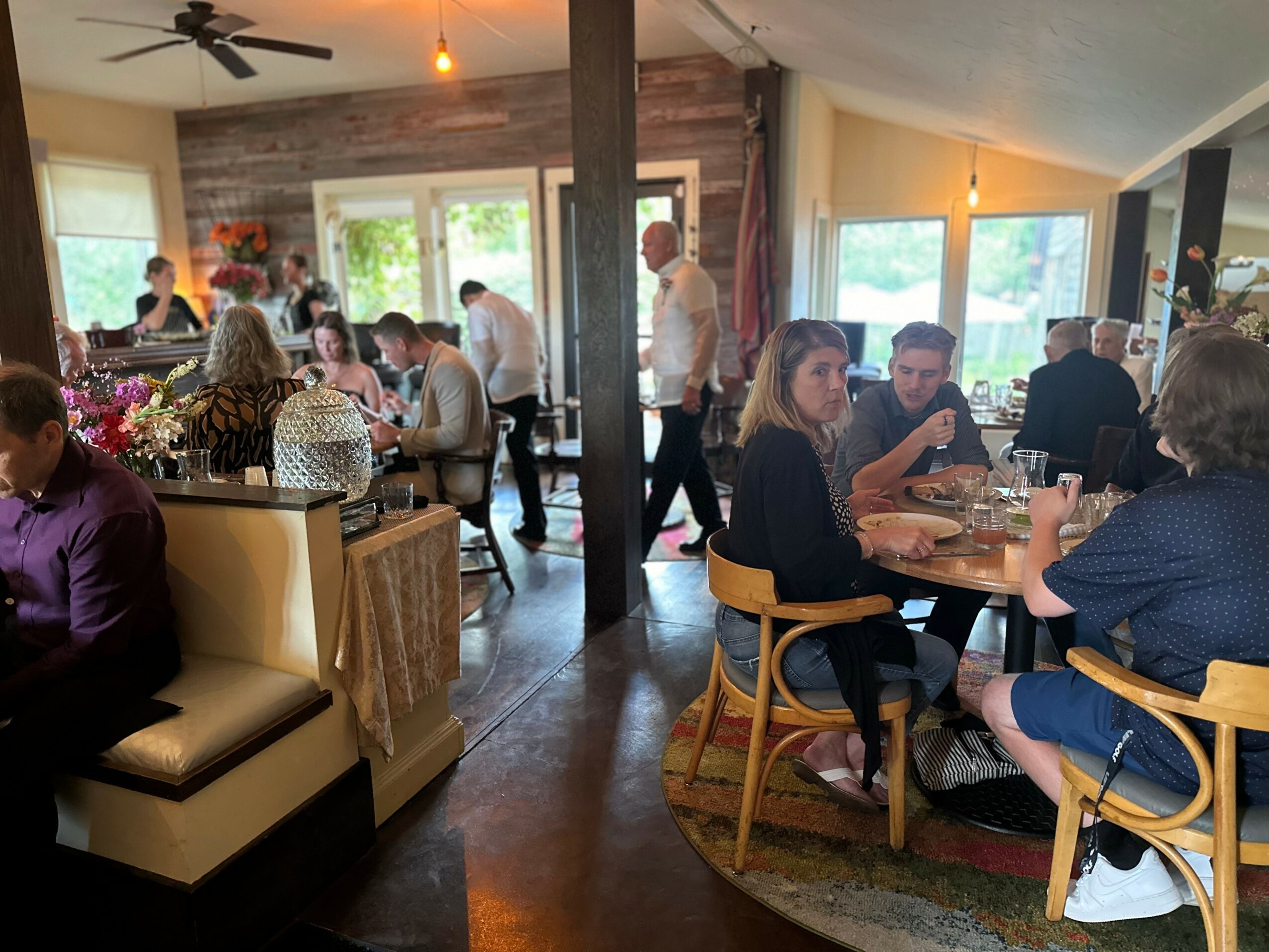 People dining in a cozy restaurant with wooden walls, large windows, and a mix of seated and standing guests.