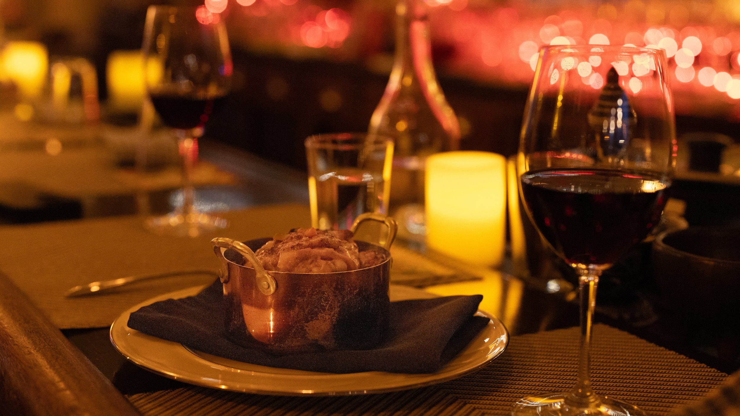 A candlelit table with a glass of red wine, a small dish, and a lit candle, in a dimly lit setting.