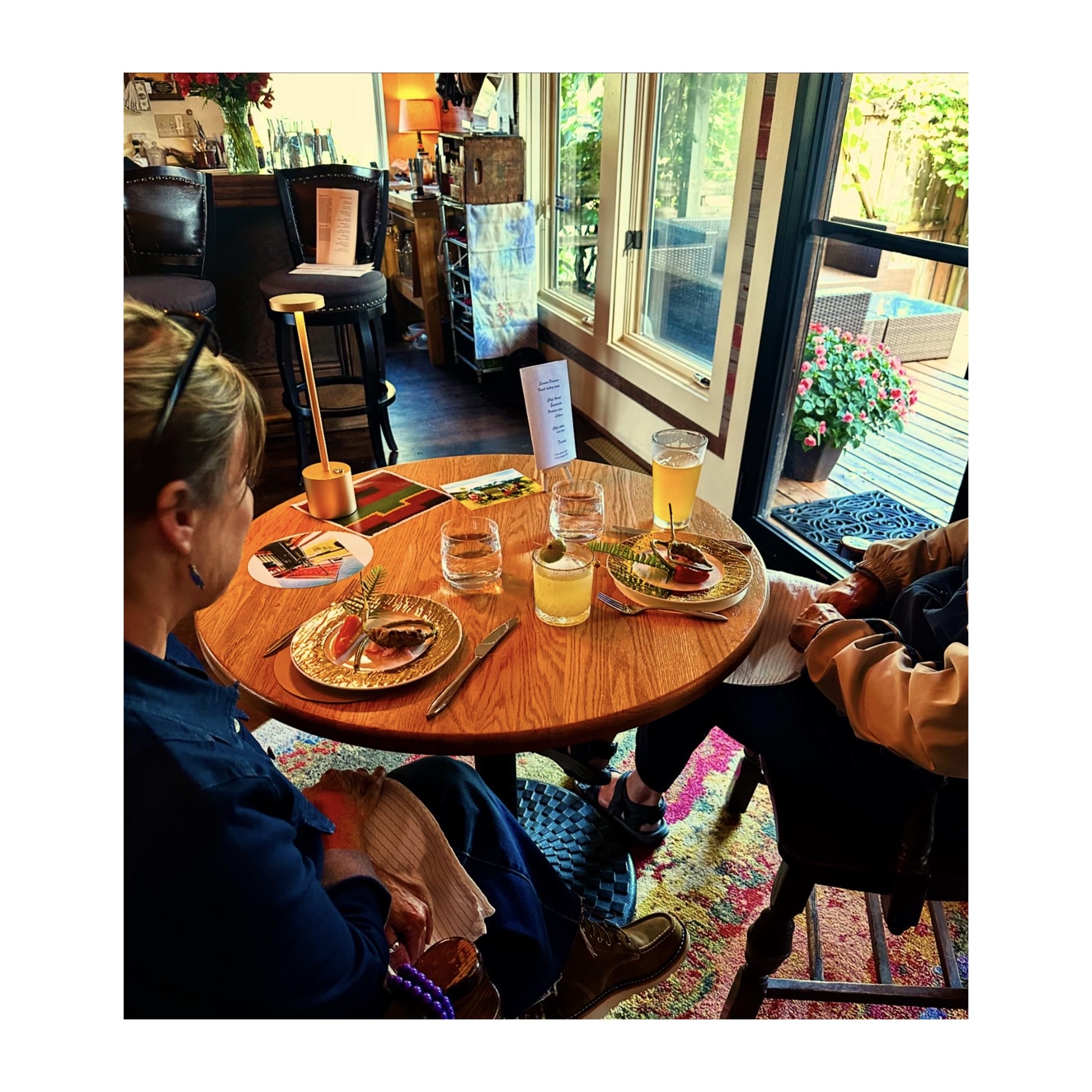 Two people sit at a round table with drinks and plates, near a window with a view of a deck and garden.