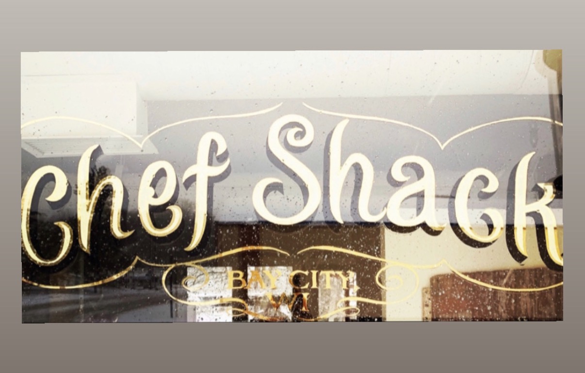 Sign with the words 'Chef Shack' in stylized font, with a background of buildings and city lights at night.