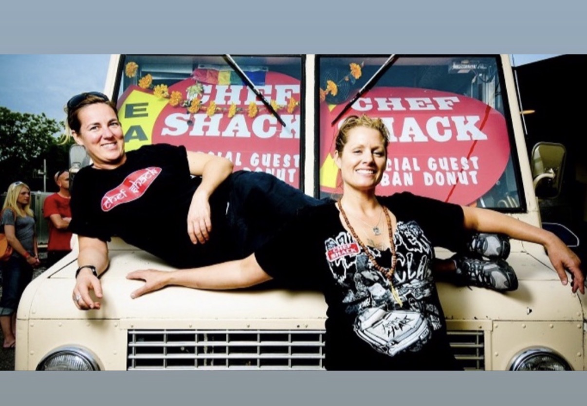 Two women lying on a vehicle in front of a sign for a food show, smiling and relaxed.