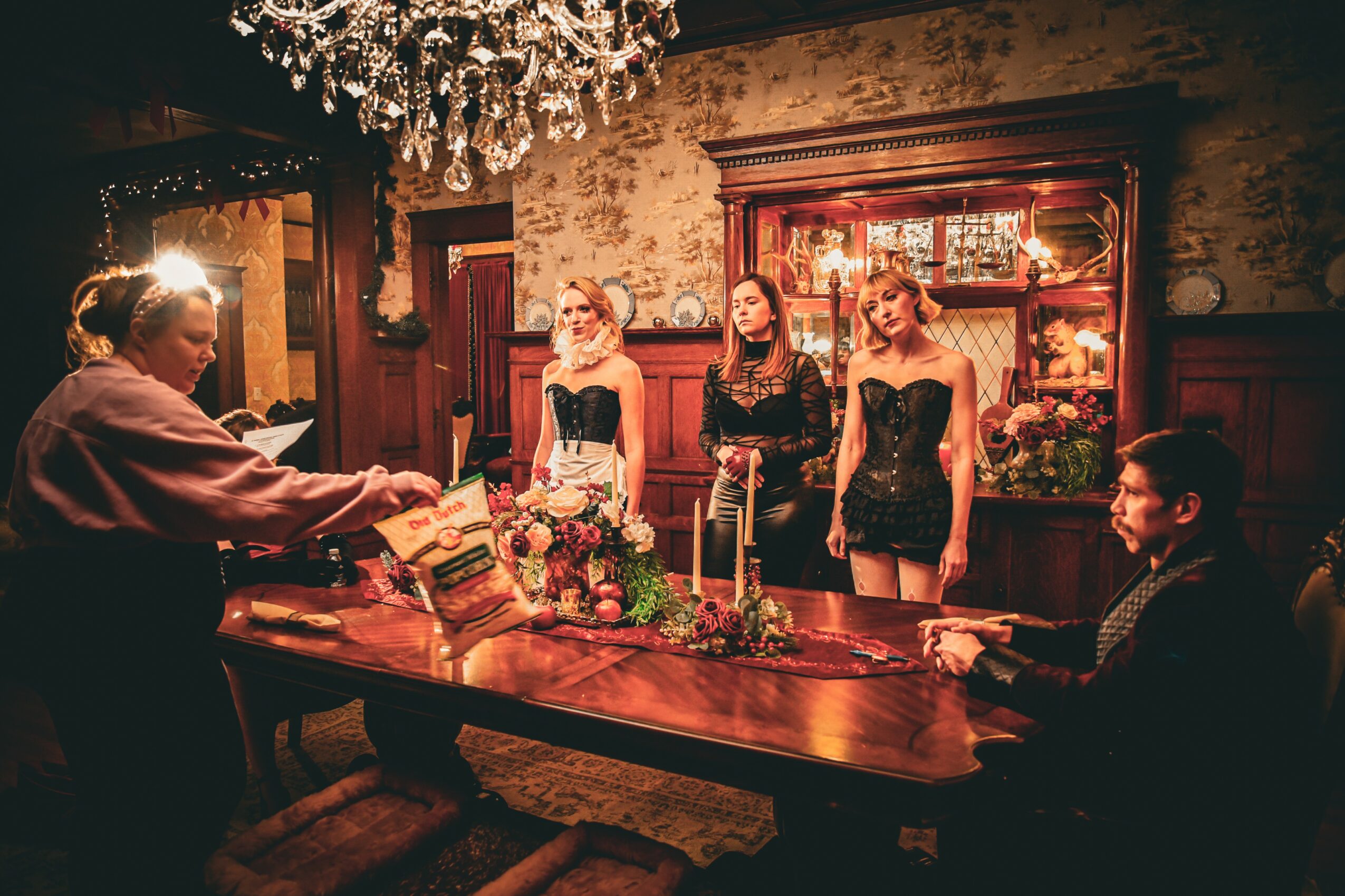 Four women stand behind a table with flowers and candles, two men sit at the table in a decorated room.