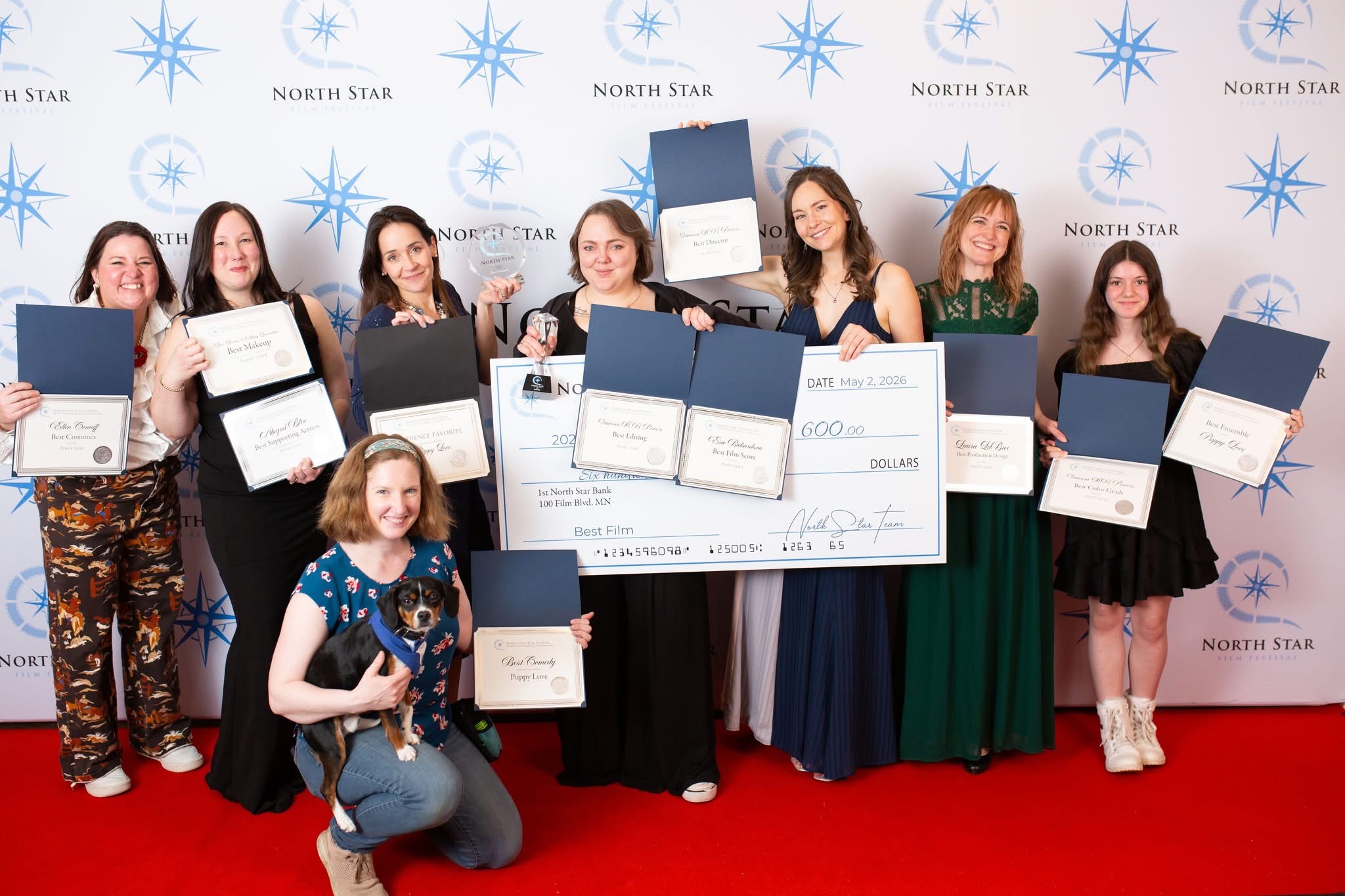 Group of nine women and one girl holding certificates and a large check, standing on a red carpet in front of a North Star backdrop.