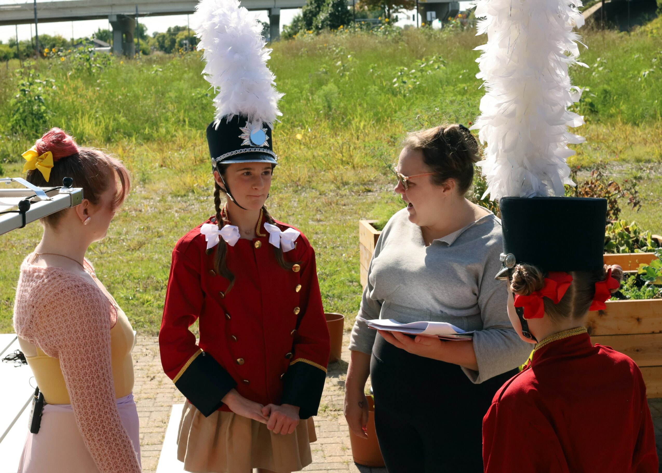 Four people outdoors, two women and two children, one dressed as a soldier, with tall feathered hats, in a grassy area.