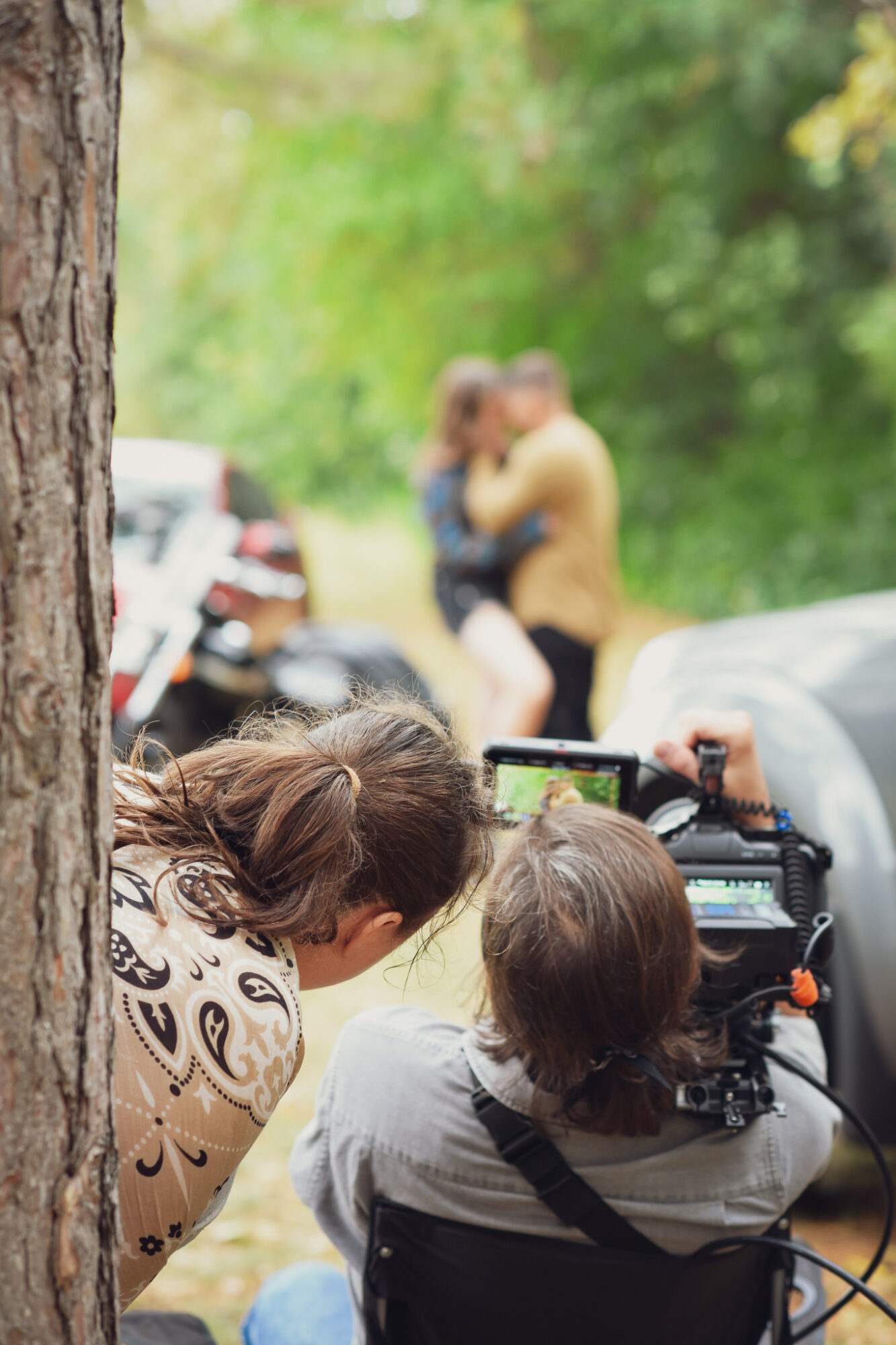 Two women are operating a camera outdoors, with a blurred couple embracing in the background among trees and parked cars.