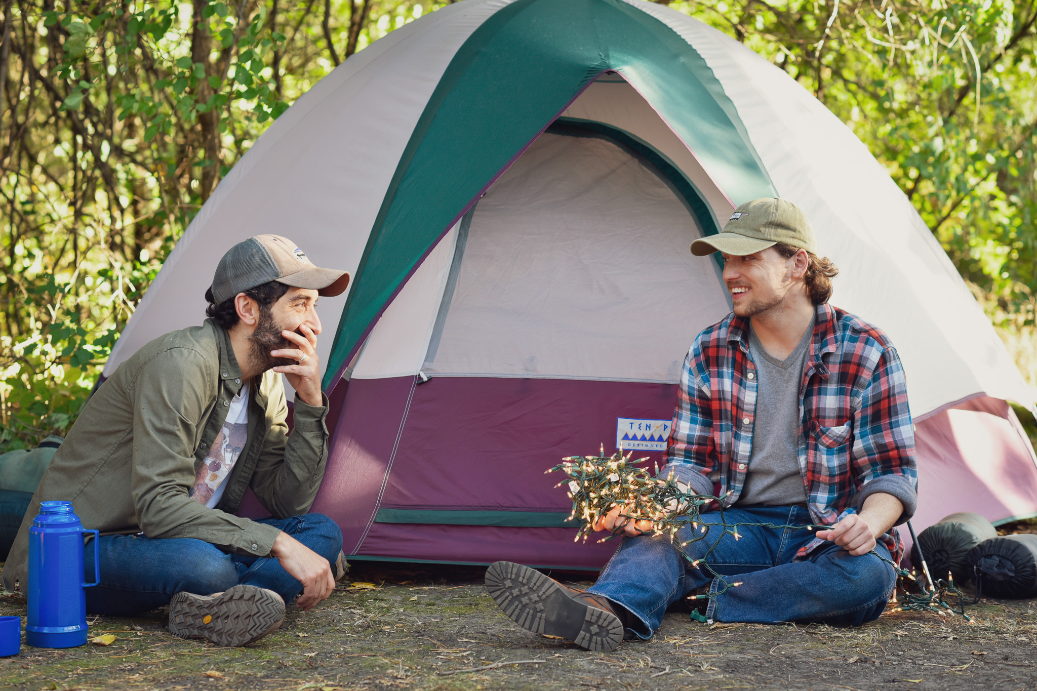 Two people sitting outside a tent in a forest, talking and smiling, with trees and sunlight in the background.