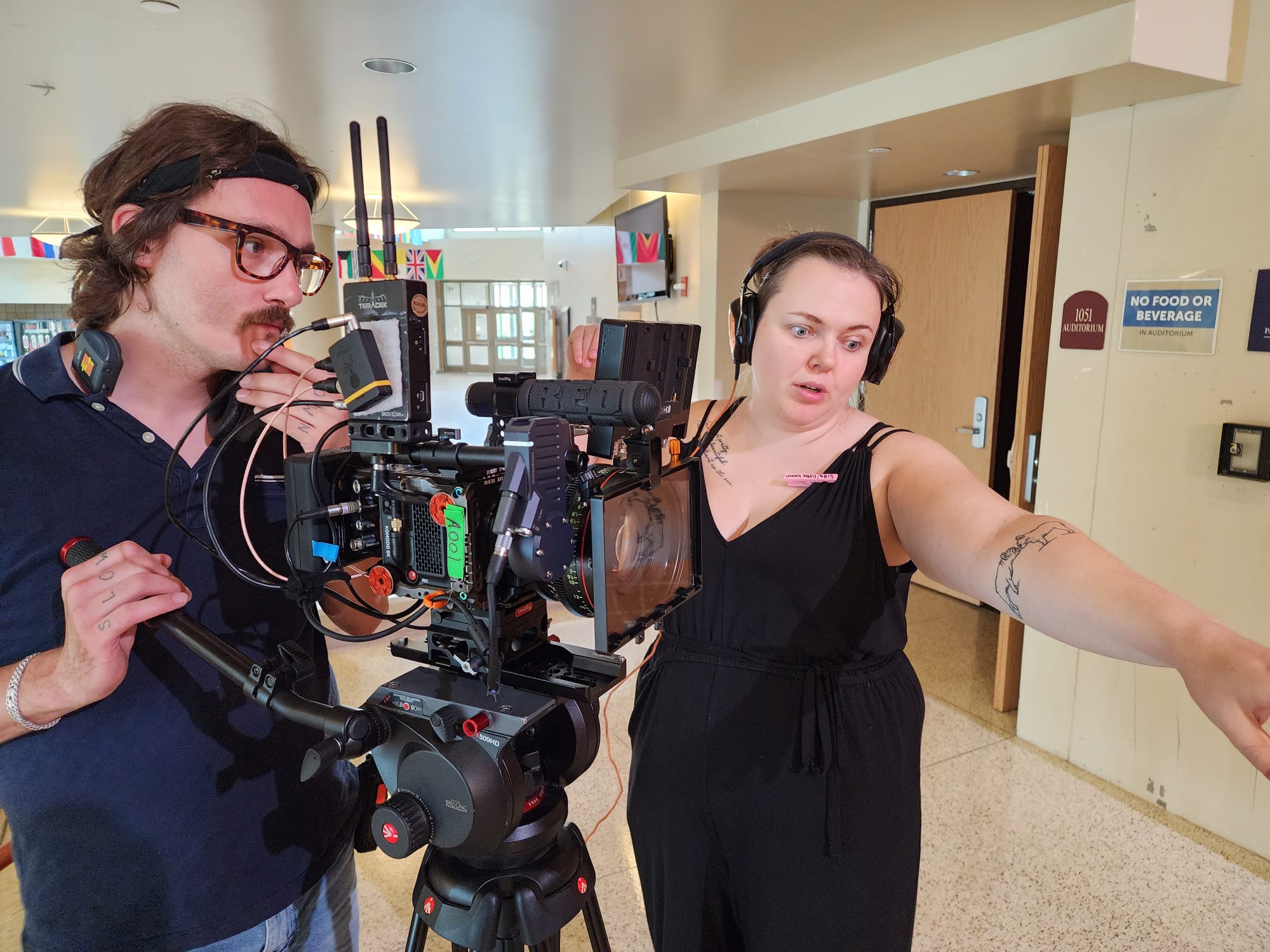 Two people operating a camera and equipment indoors, one woman pointing, the other man adjusting controls.