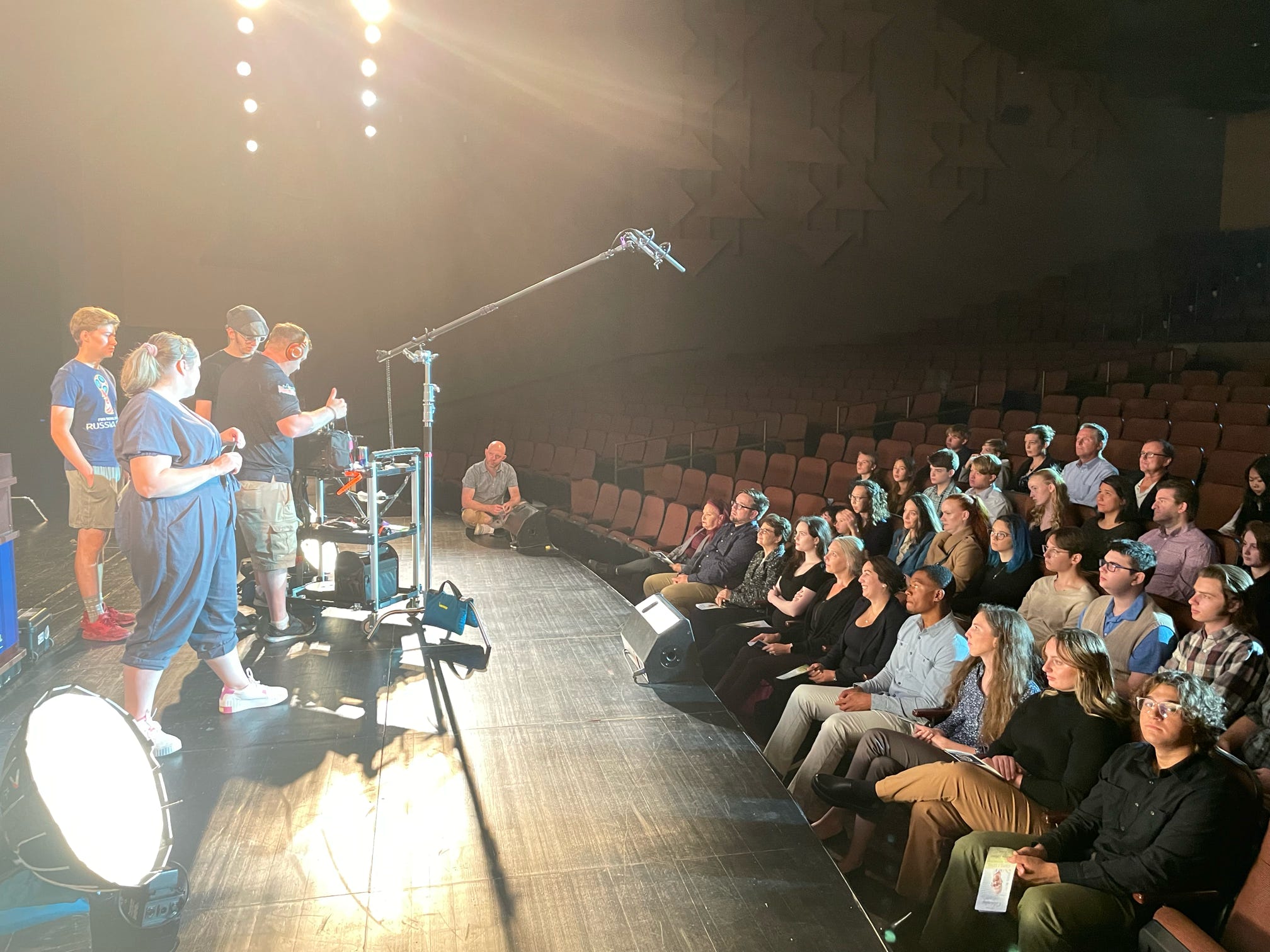 People on stage performing for seated audience in a theater with empty seats, stage lighting, and camera equipment.