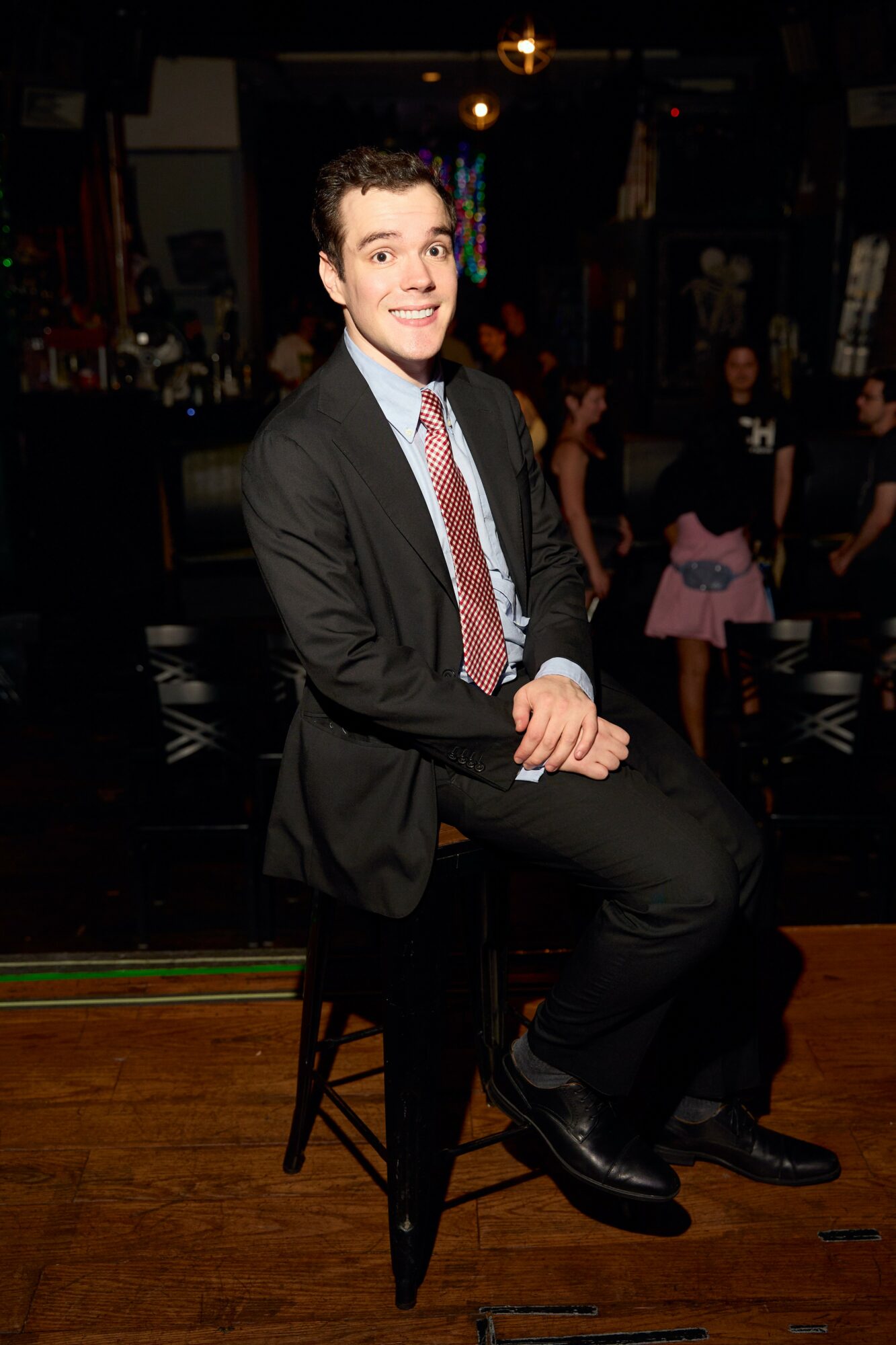 Young man in a suit sitting on a stool, smiling, in a dimly lit social setting with people in the background.