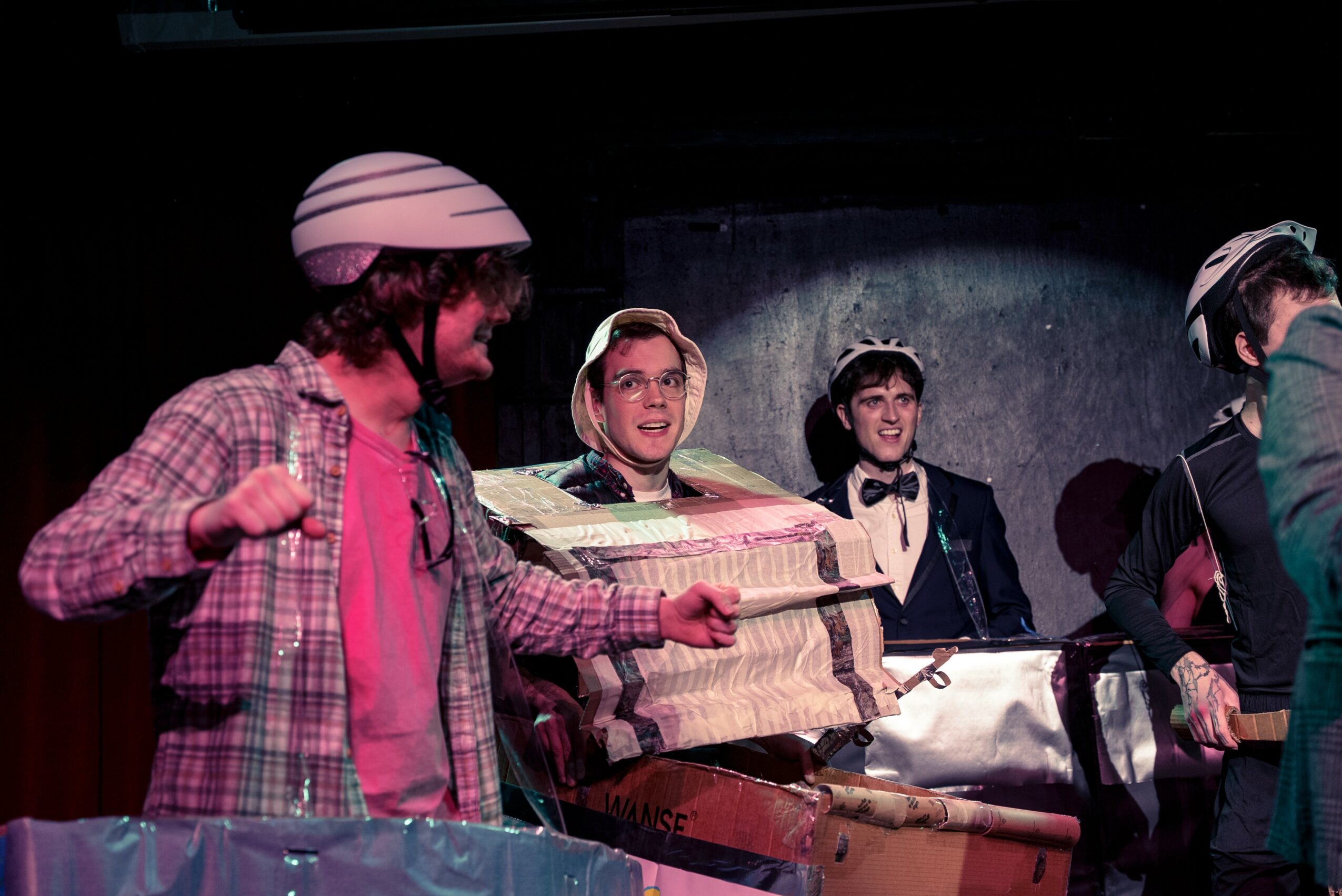 Four people on stage, one holding a large box, with a dark background and stage lighting.