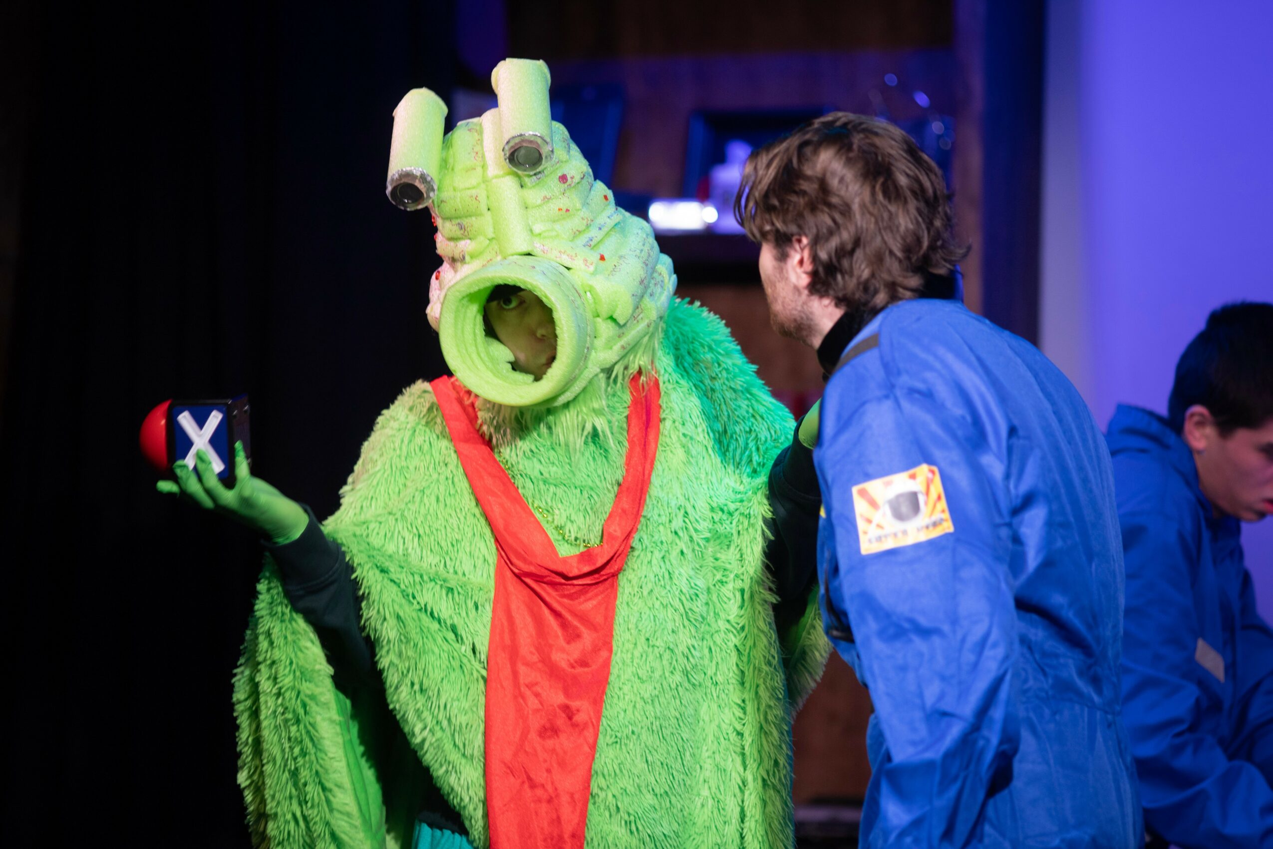 Person in a green costume with a large headpiece talking to a person in a blue jacket, stage background.