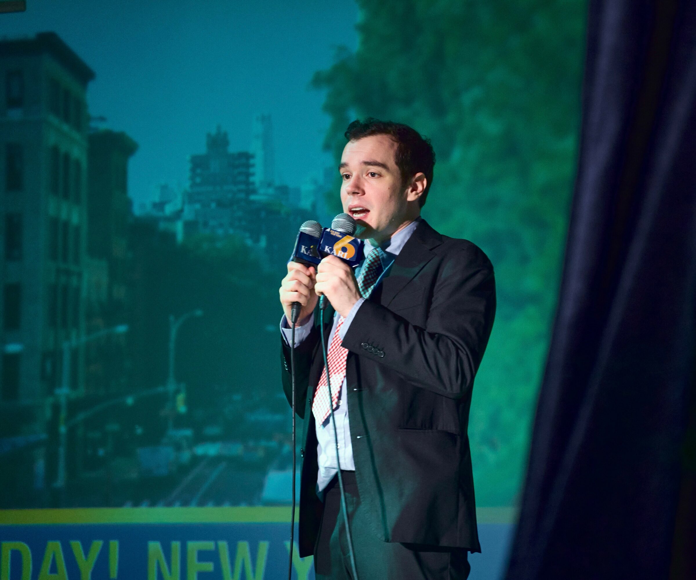 Young man in suit singing into a microphone on stage with cityscape background.