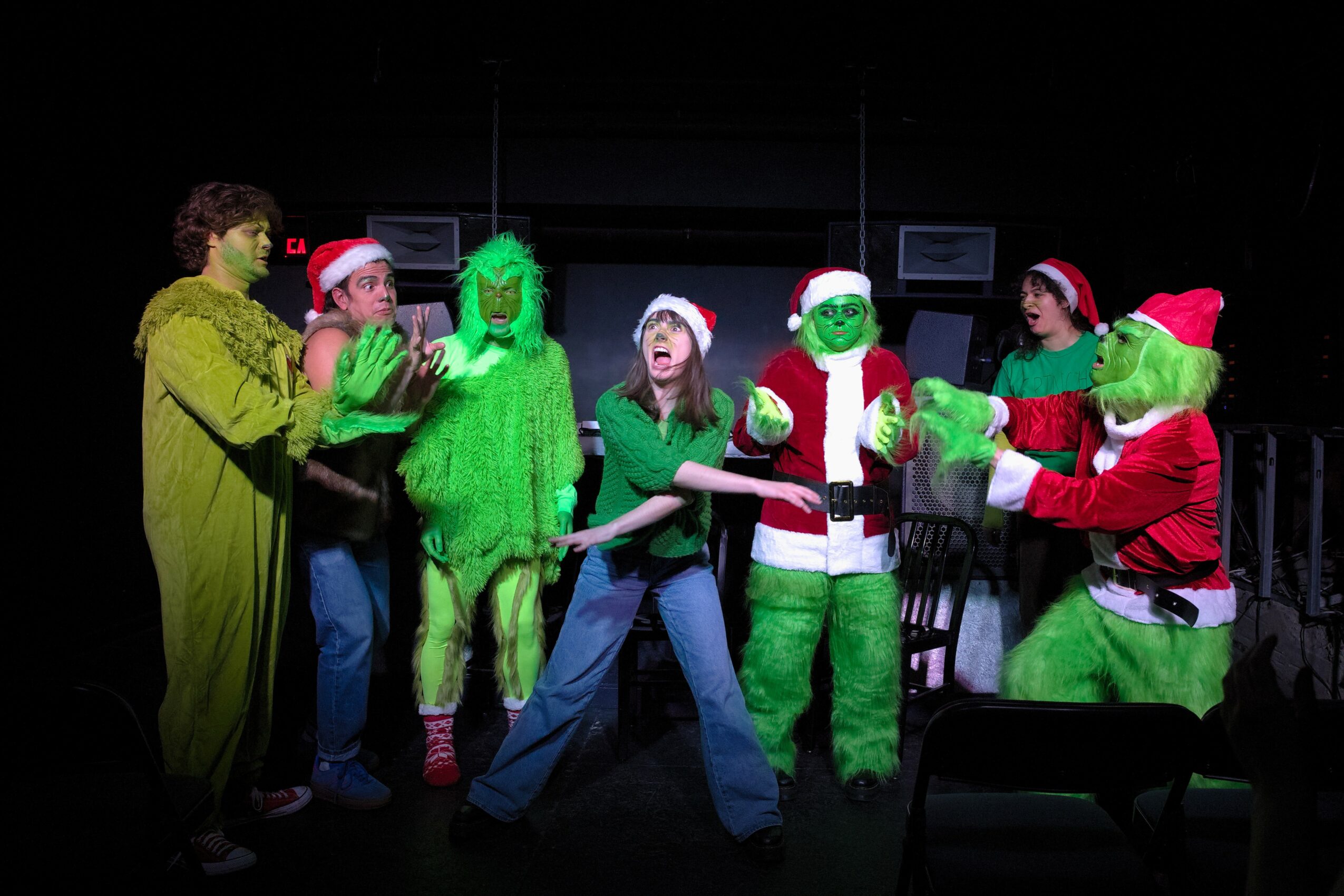 People in Christmas costumes, including Grinch characters, gather on stage with black background, some wearing Santa hats, engaging in a performance.