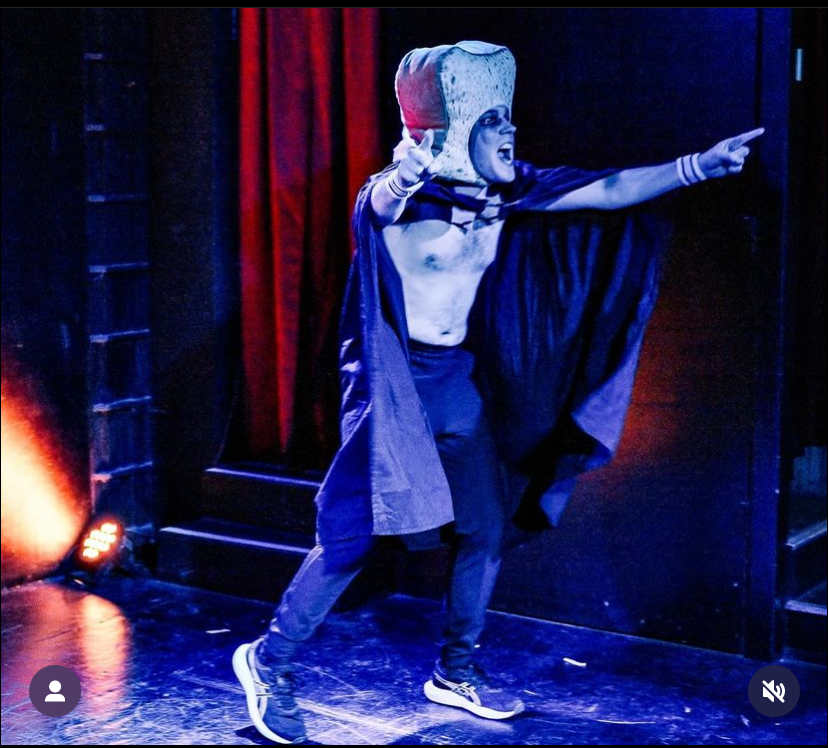 Child in a costume with a tall headpiece, wearing a cape, on stage with red curtains in background.