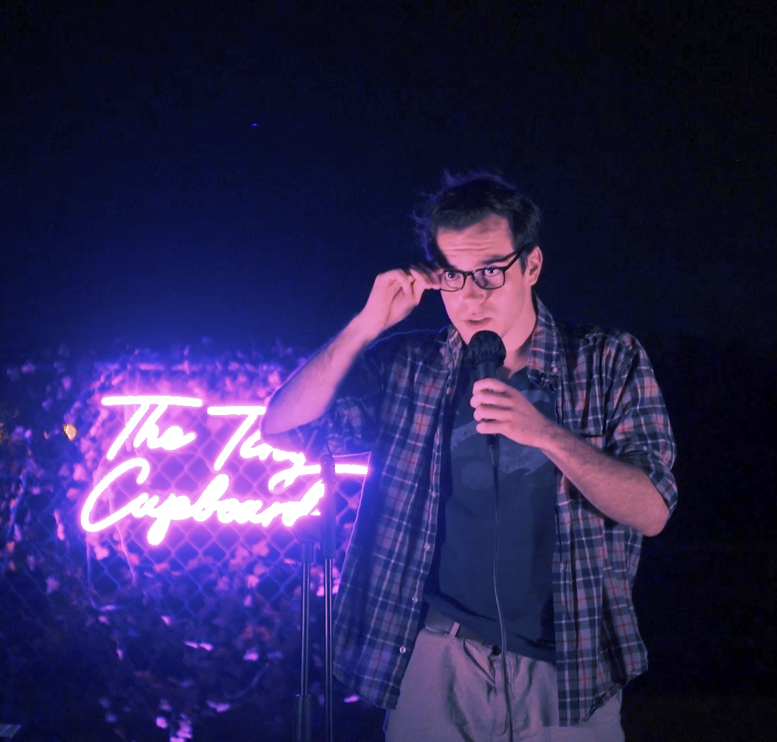 Person with glasses holding microphone, adjusting glasses, standing in front of neon sign, dark background, casual clothing.