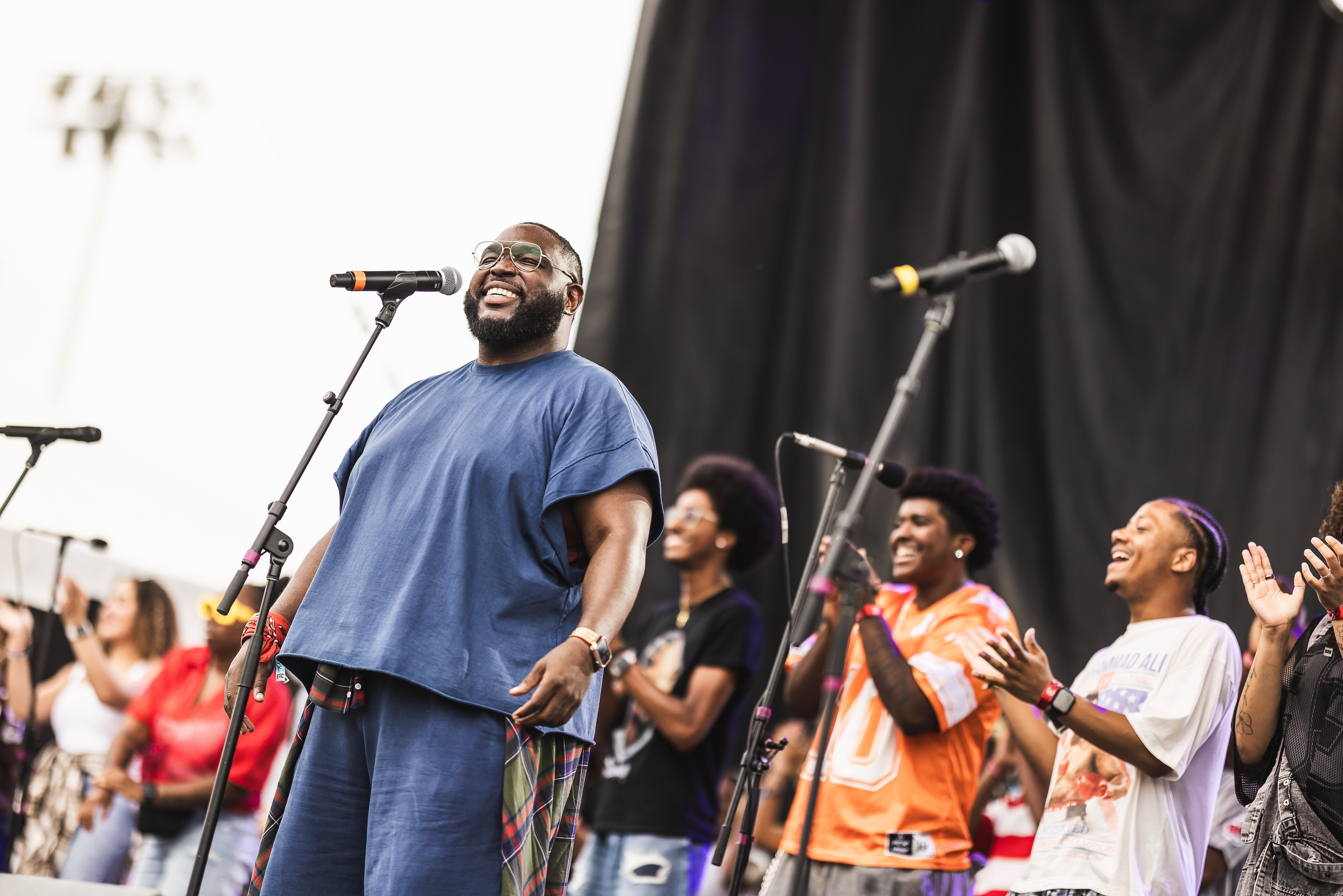 Man with glasses and beard smiling on stage, surrounded by people clapping and enjoying performance.