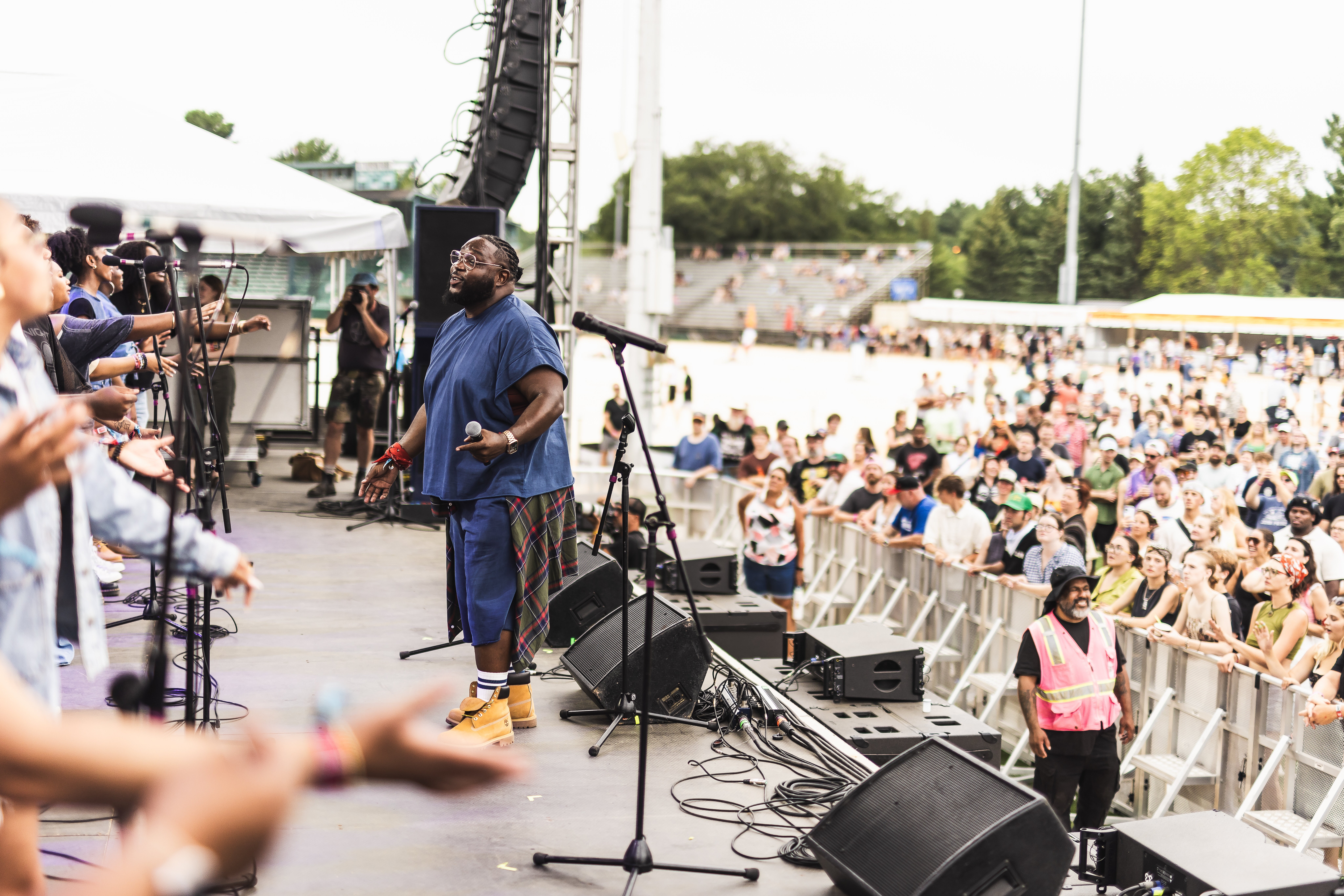 Performer on stage with microphone, audience watching outdoors, security guard in pink vest, many people seated, trees in background.