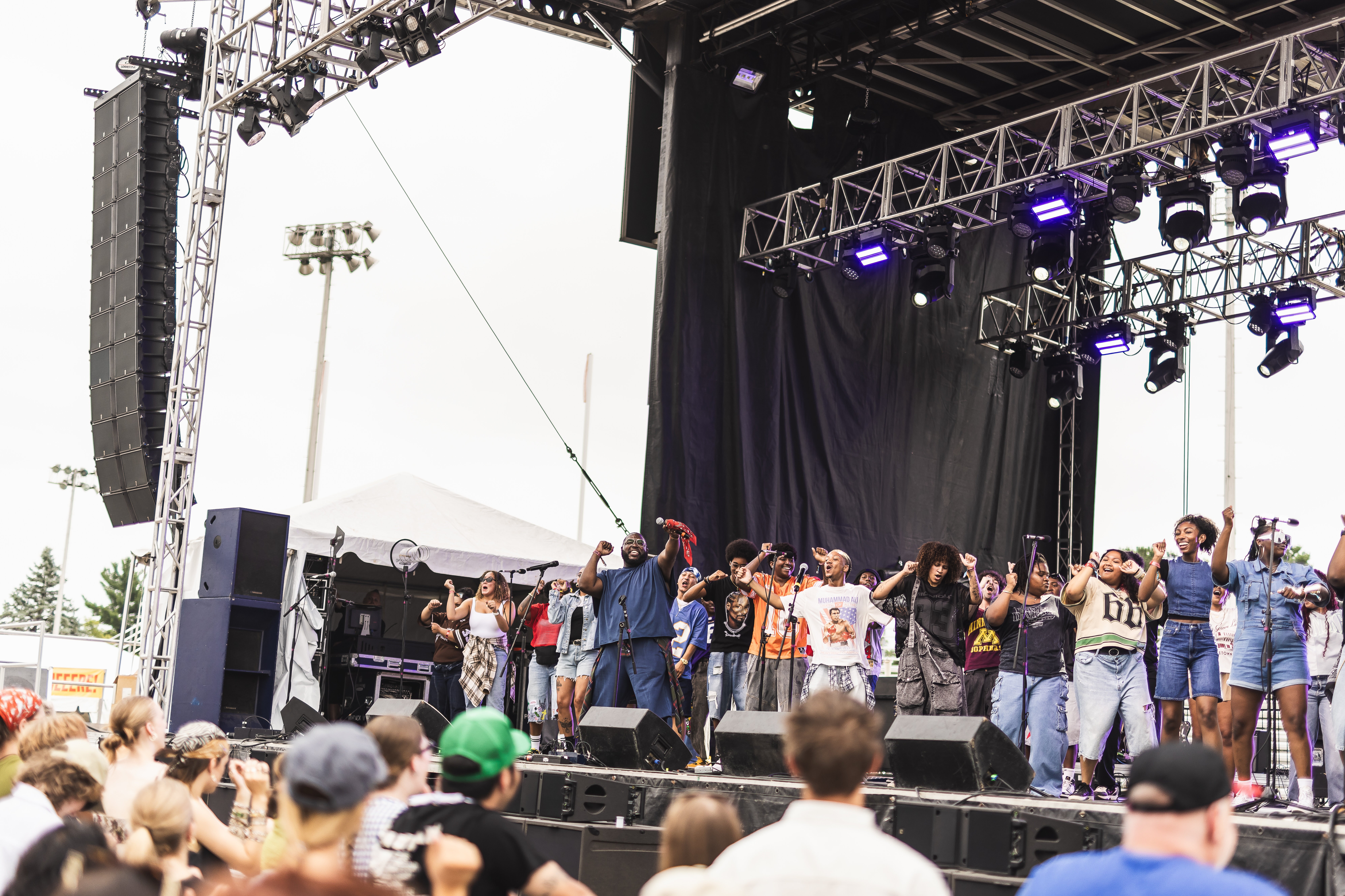 Group of people performing on an outdoor stage with lighting and sound equipment, audience watching, some taking photos.