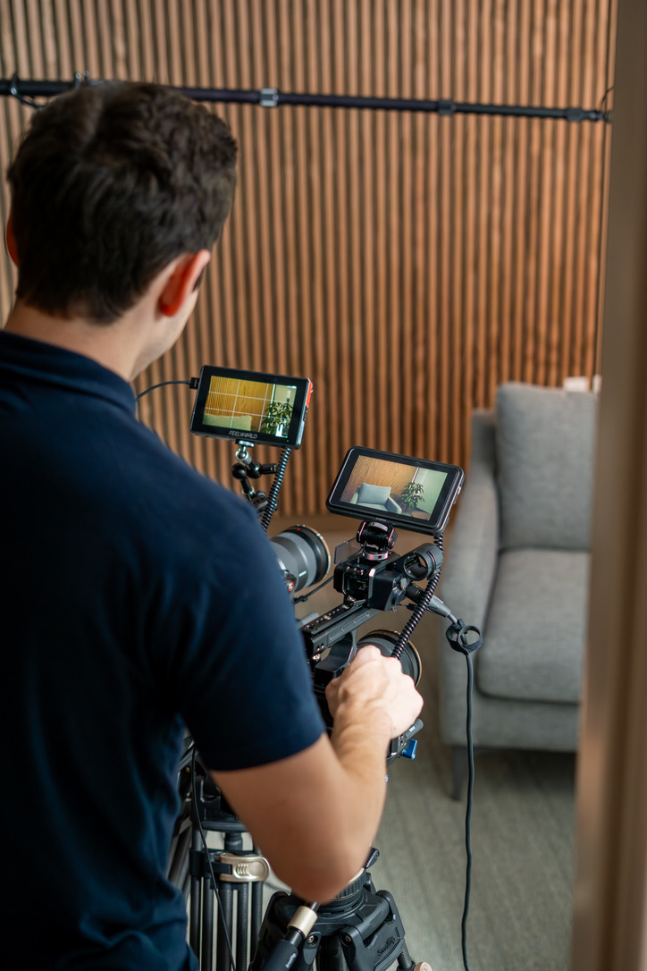 Person operating a camera with two monitors, facing a wooden wall and a gray chair in a room.