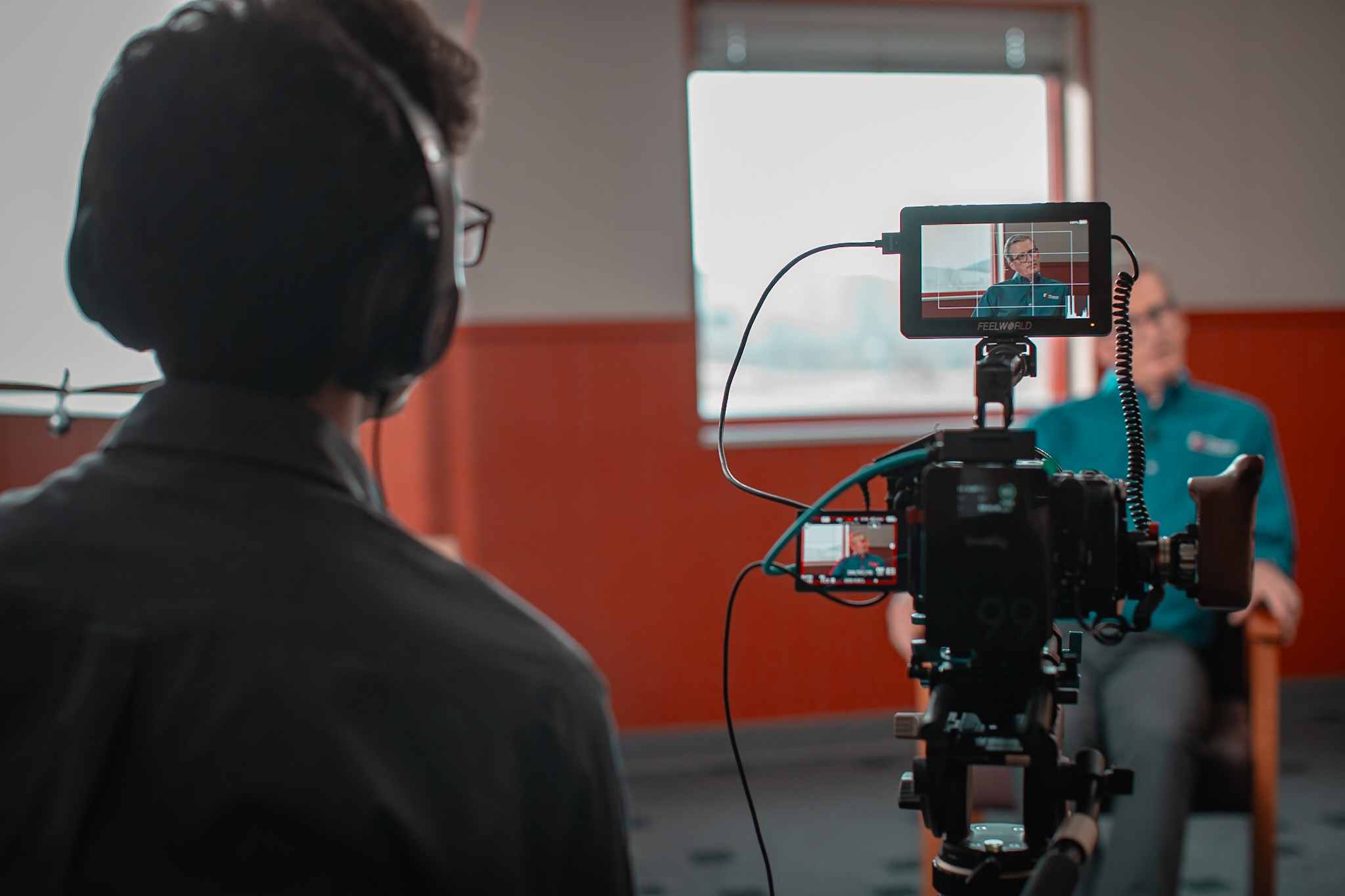 Person with headphones filming a woman sitting on a chair in a room with a window and orange wall.
