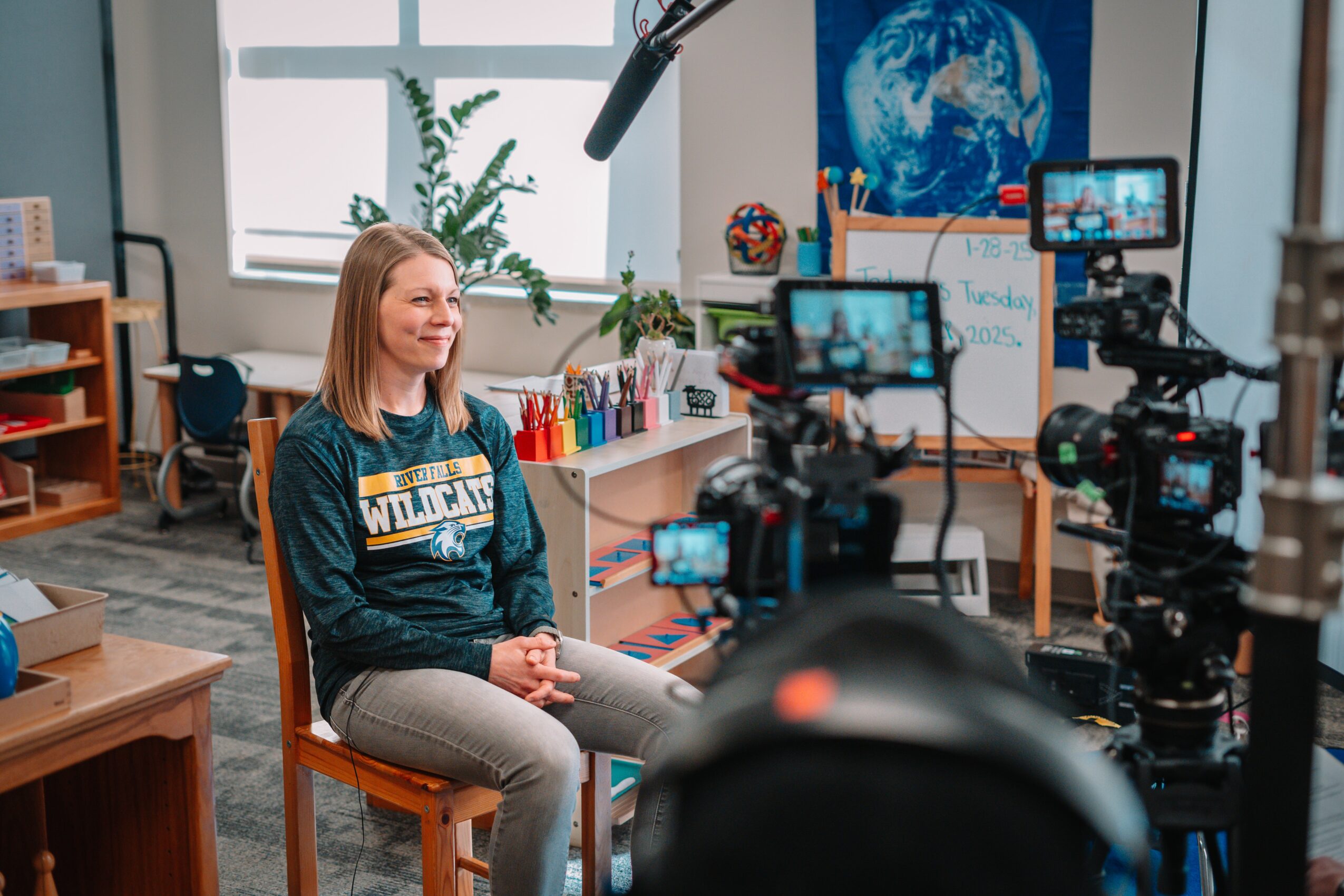 Young woman sitting in a classroom, being filmed with professional cameras, with a globe and educational posters in the background.