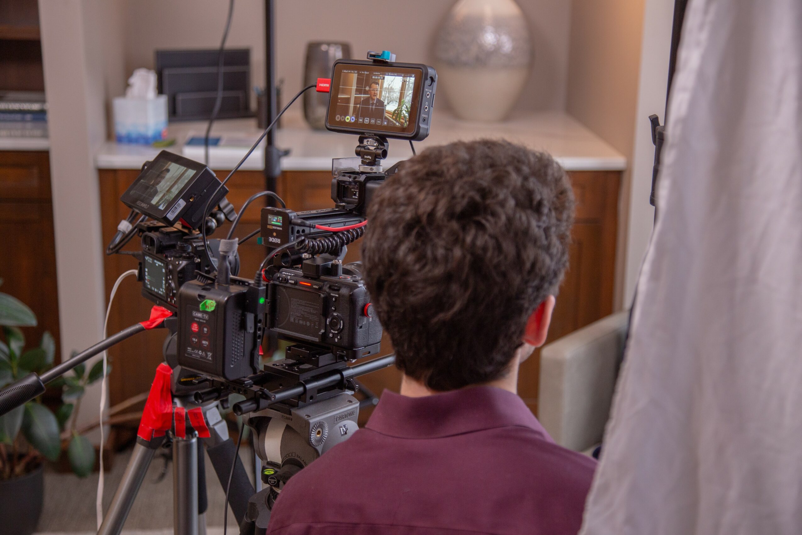 Person with curly hair seen from behind, filming with professional camera equipment indoors, with a white curtain on the right.
