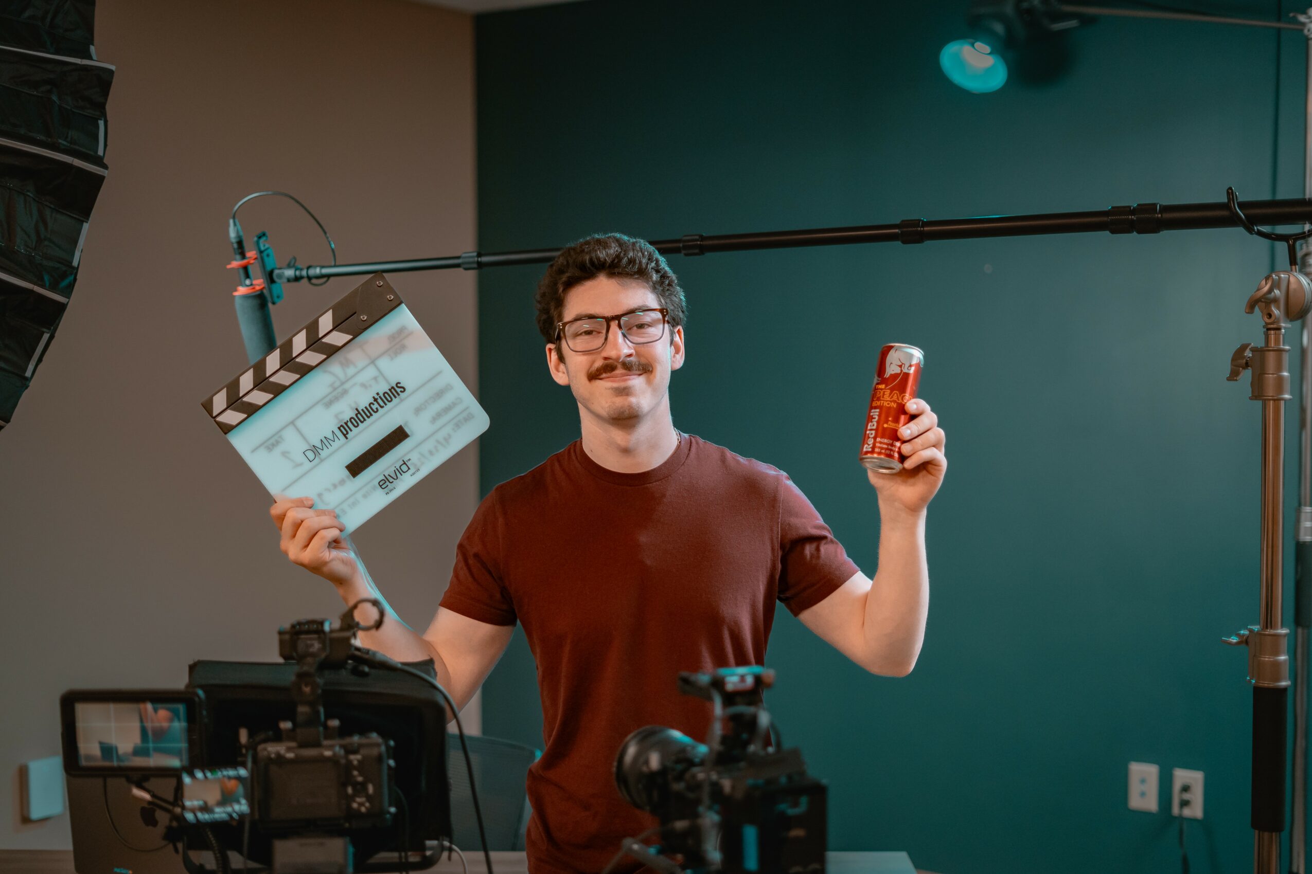 Young man with glasses holding a film clapperboard in one hand and a drink in the other, smiling in a studio.