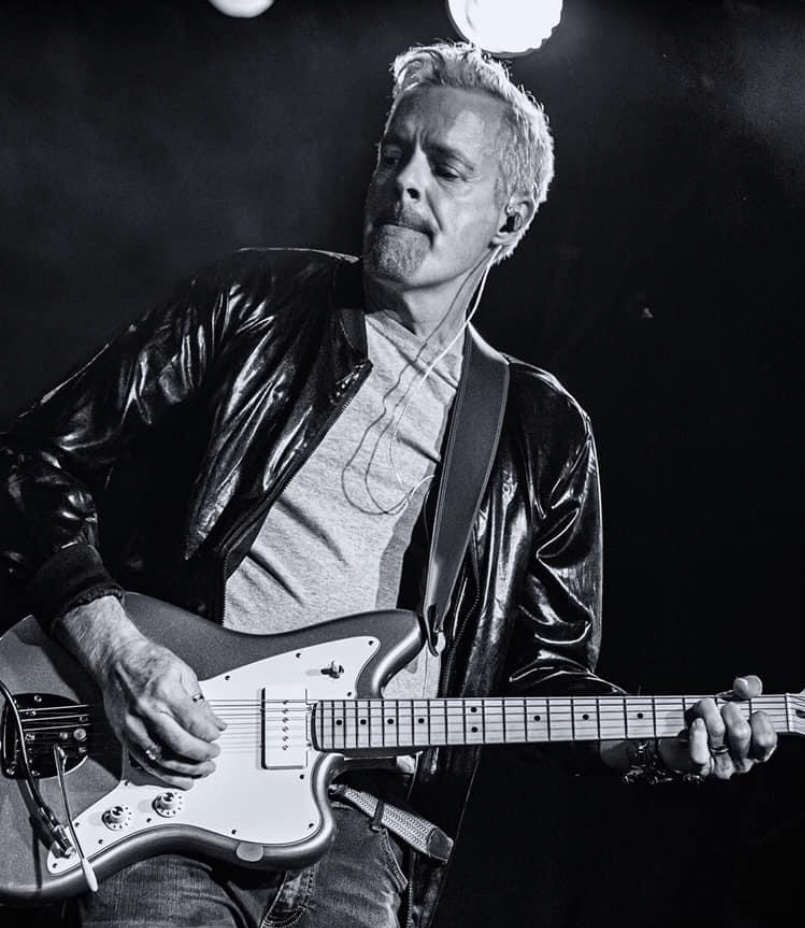 Man playing electric guitar, wearing a leather jacket, with a focused expression, in black and white.