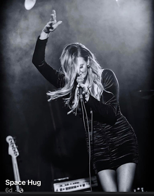 Female singer performing on stage with microphone, hair covering face, one arm raised, in black and white.