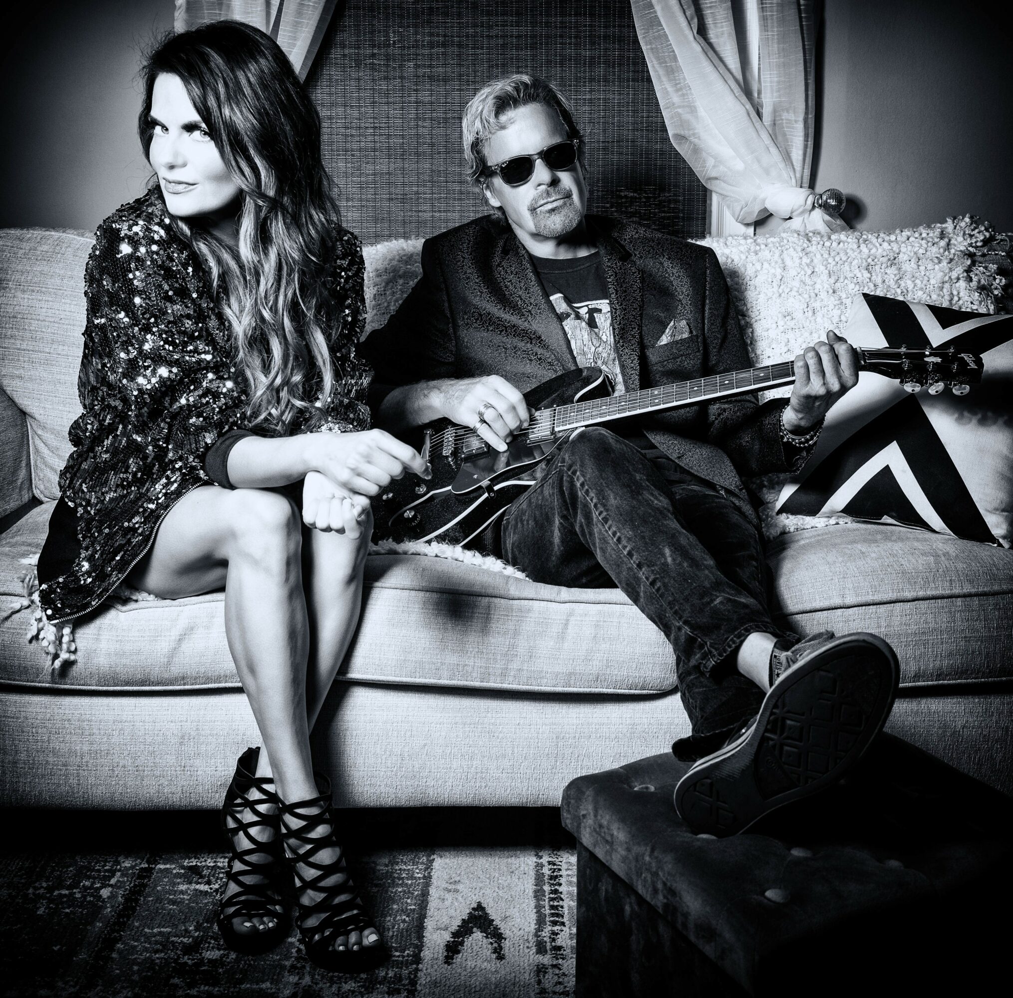 Two people sitting on a sofa, one holding a guitar, in a room with curtains and a patterned rug, black and white.