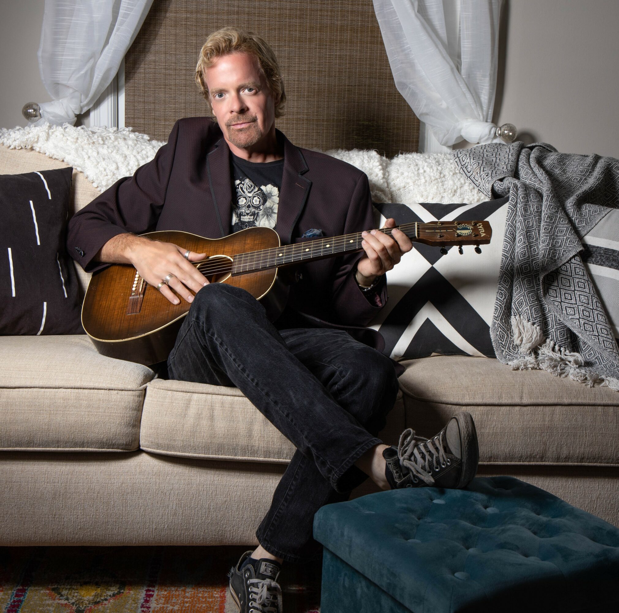 Man sitting on a sofa holding an acoustic guitar, with curtains and blankets in the background.